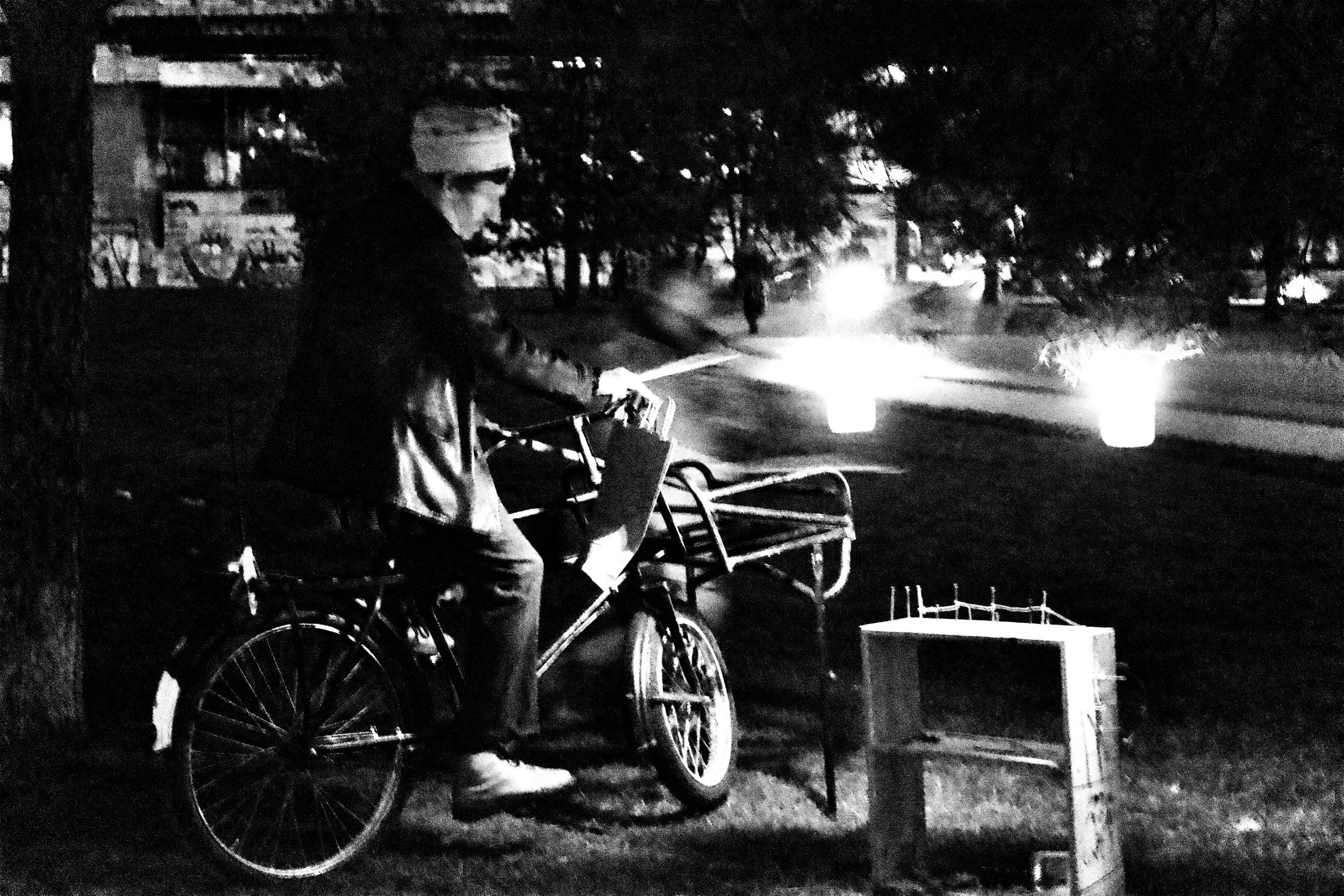 A person standing next to a bicycle at night, wearing a beanie and hoodie. They are beside dispensing containers and a small cart, with trees and streetlights in the background.