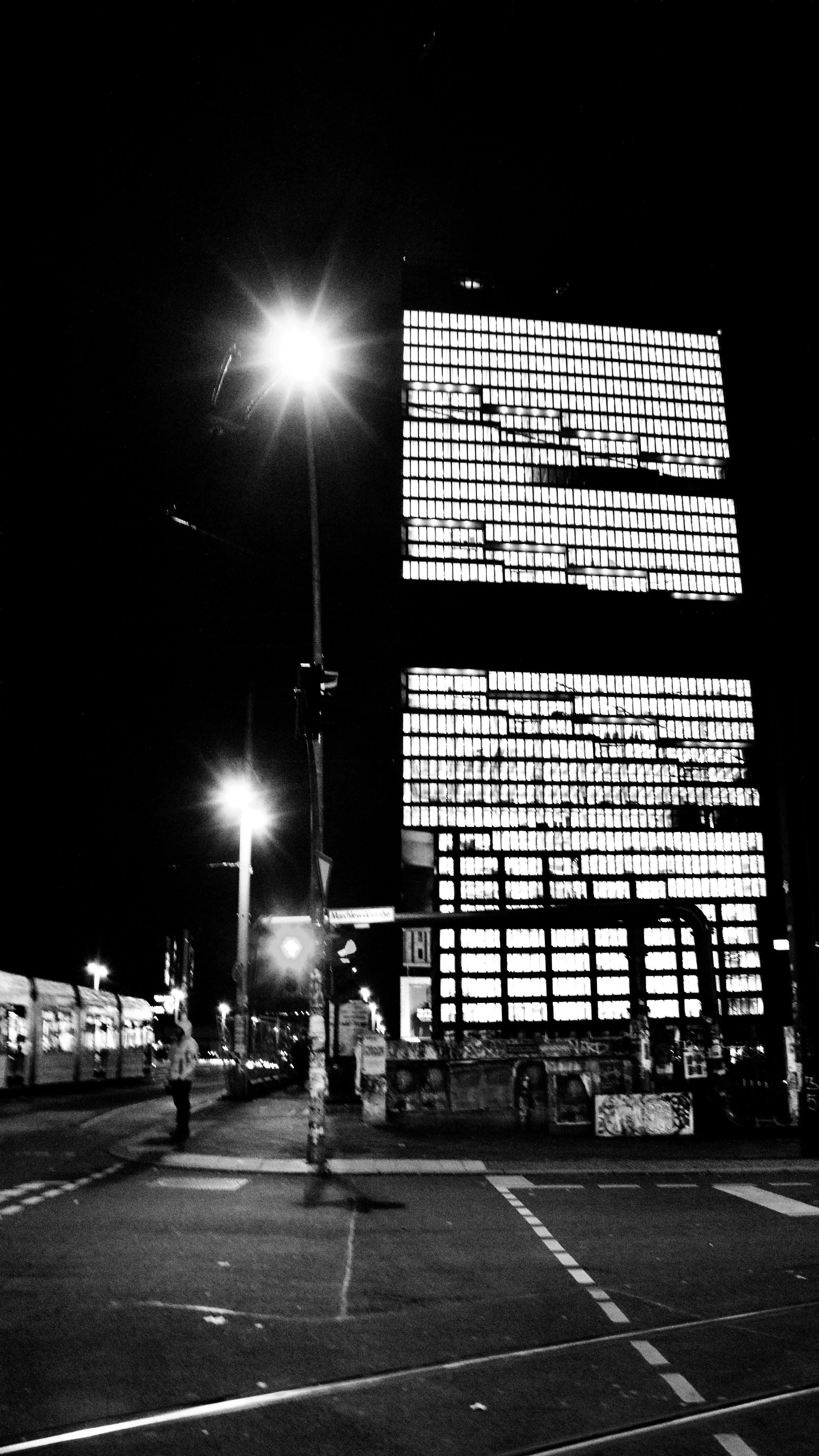 Night scene of a city street with a tall building with illuminated windows, street lamps, and few people walking on the sidewalk.