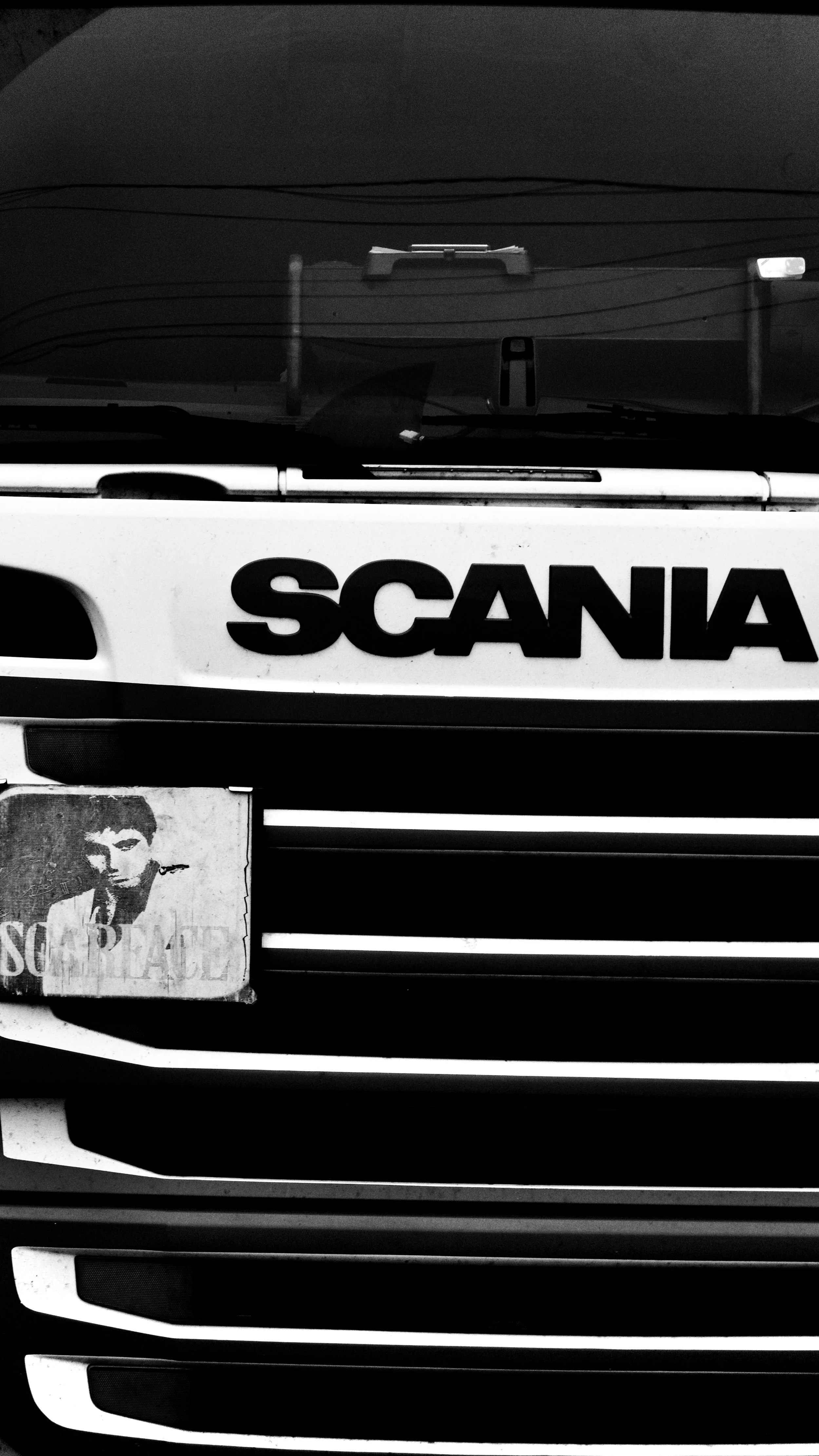 Black and white street scene featuring a vehicle with the word 'SCANIA' on the front and a poster of a young man on the lower left side.