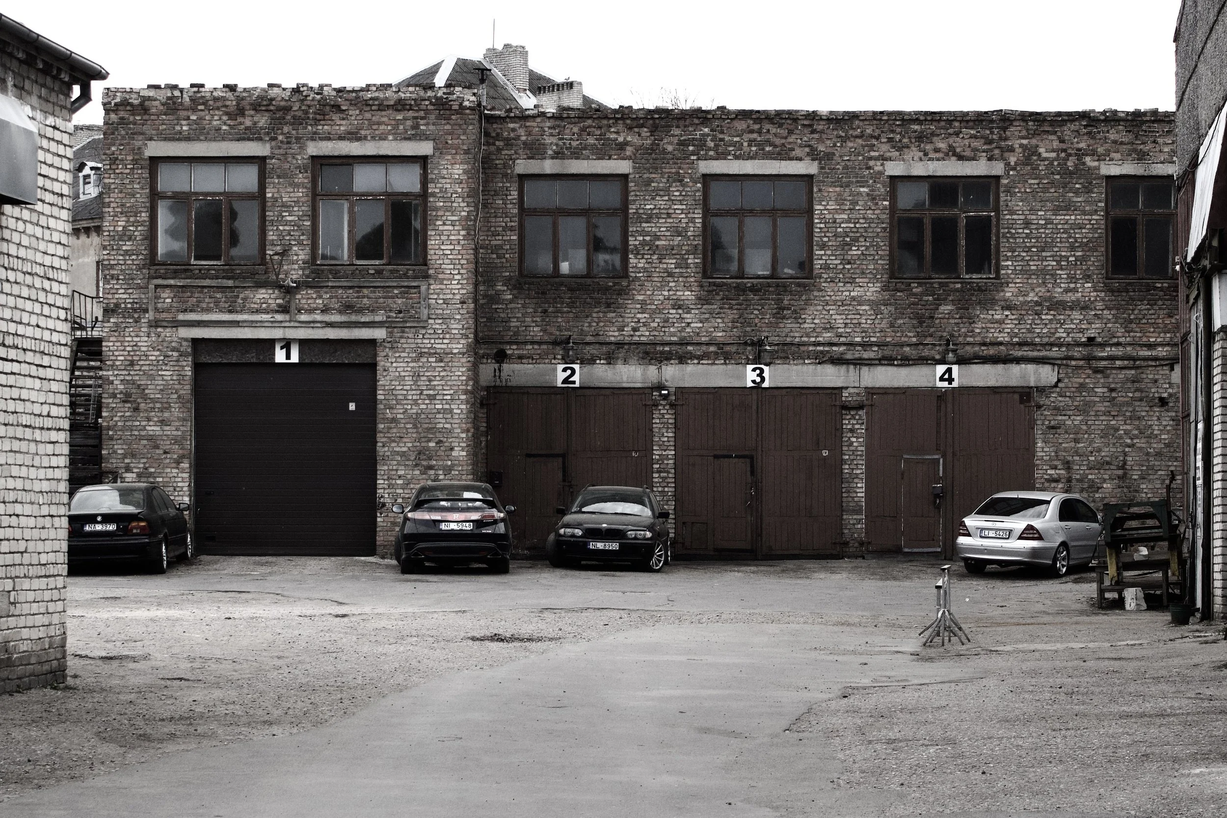 A brick building with five garage doors numbered 1 to 4 at the bottom, four parked cars in front, and three windows on top, with a cloudy sky background.