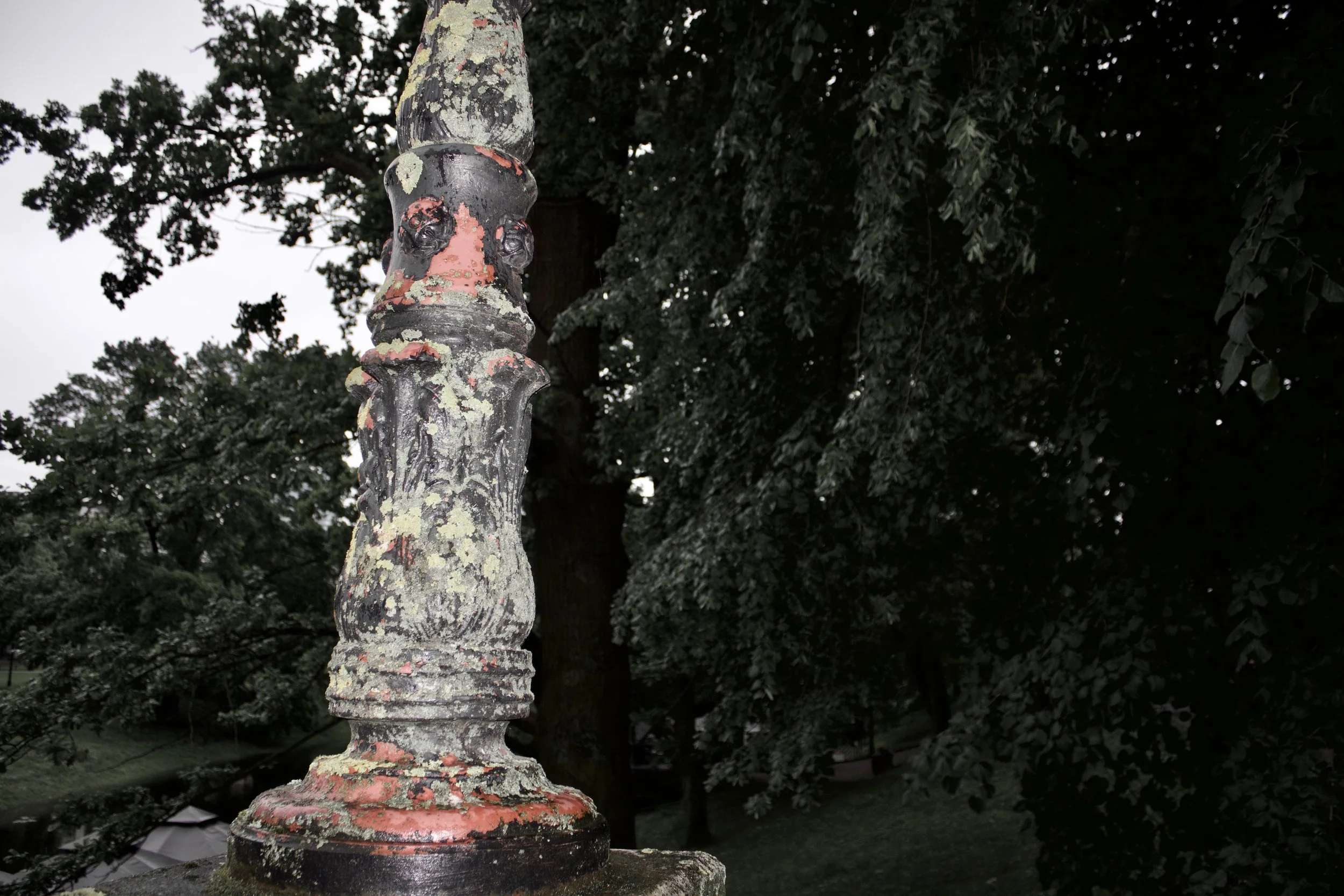 Close-up of an old, weathered, black and multicolored painted decorative metal post with peeling paint, set against a background of dense green foliage and trees.