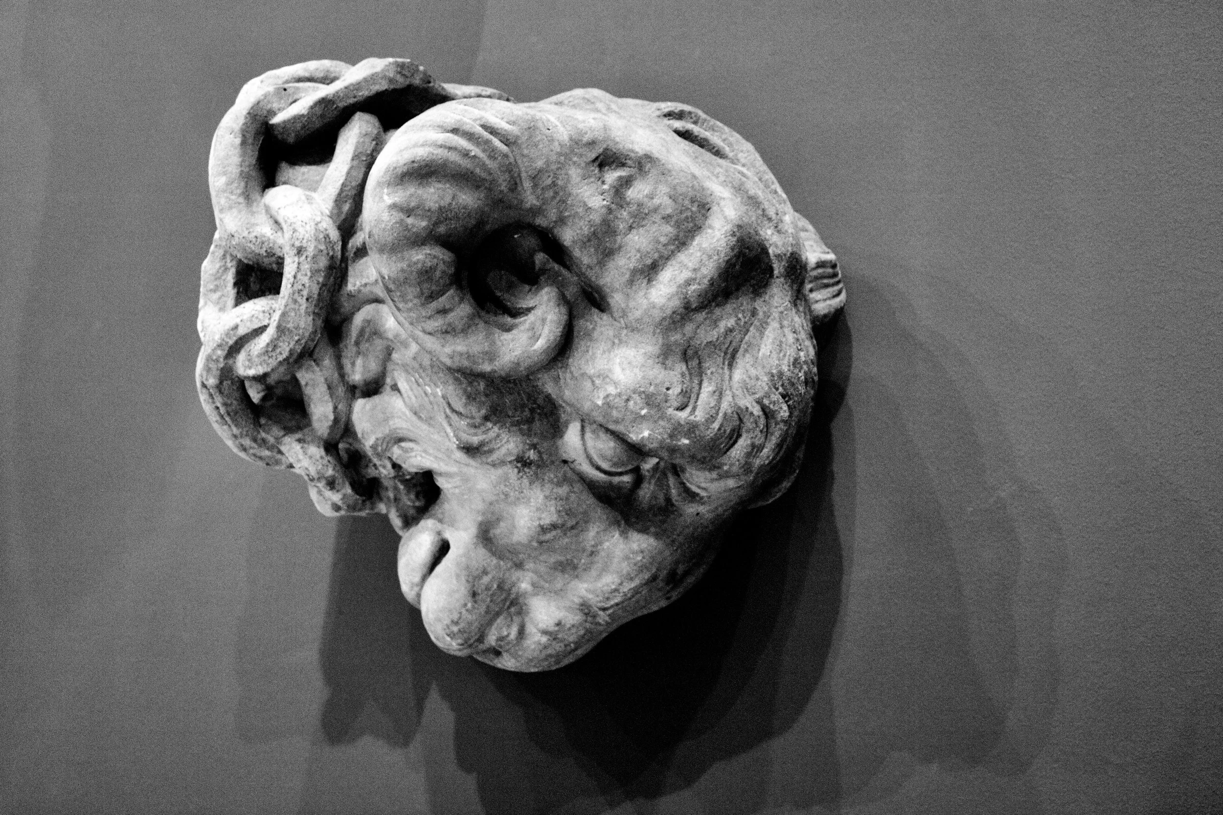 Black and white photograph of an ancient stone sculpture of a human face with detailed curly hair and prominent facial features, against a plain background.