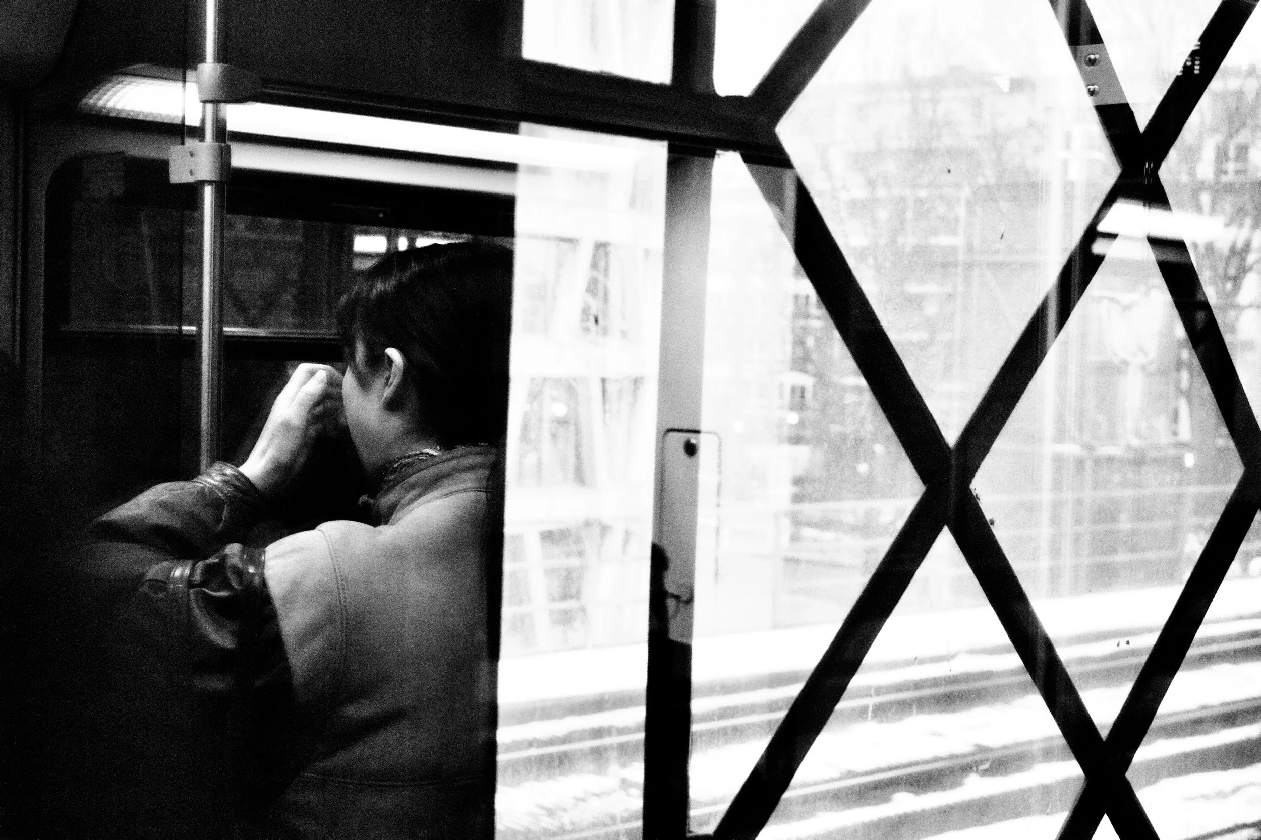 A person with dark hair, seen from behind, sitting in a room with a large window. The person appears to be listening or thinking, with their hand near their ear. The window shows an urban street scene with a building. The photo is in black and white,