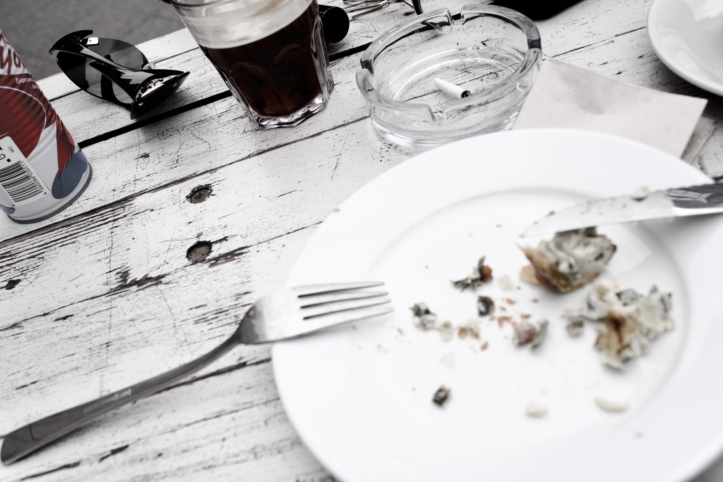 A partially eaten slice of pizza on a white plate, with crumbs and toppings visible, on a rustic wooden table. There is a fork beside the plate, an ashtray with a cigarette, a glass of dark beverage, and a can of soda, with sunglasses nearby.