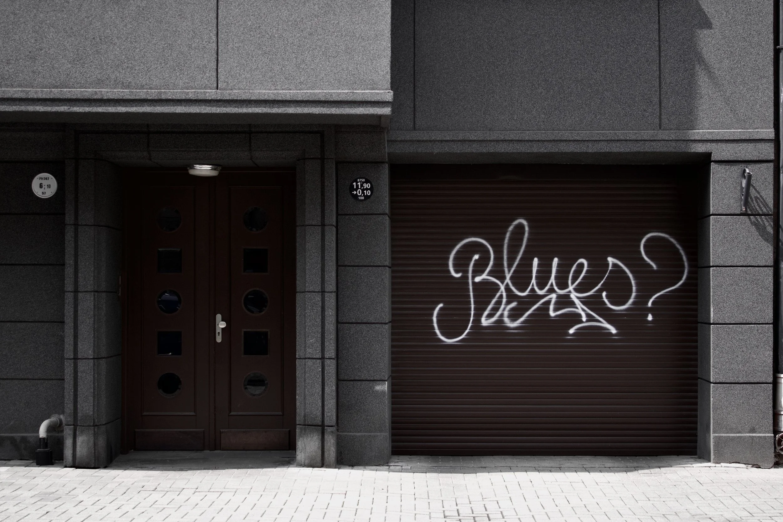 Gray building with a closed brown metal roll-up door with graffiti and a dark brown door with round windows.