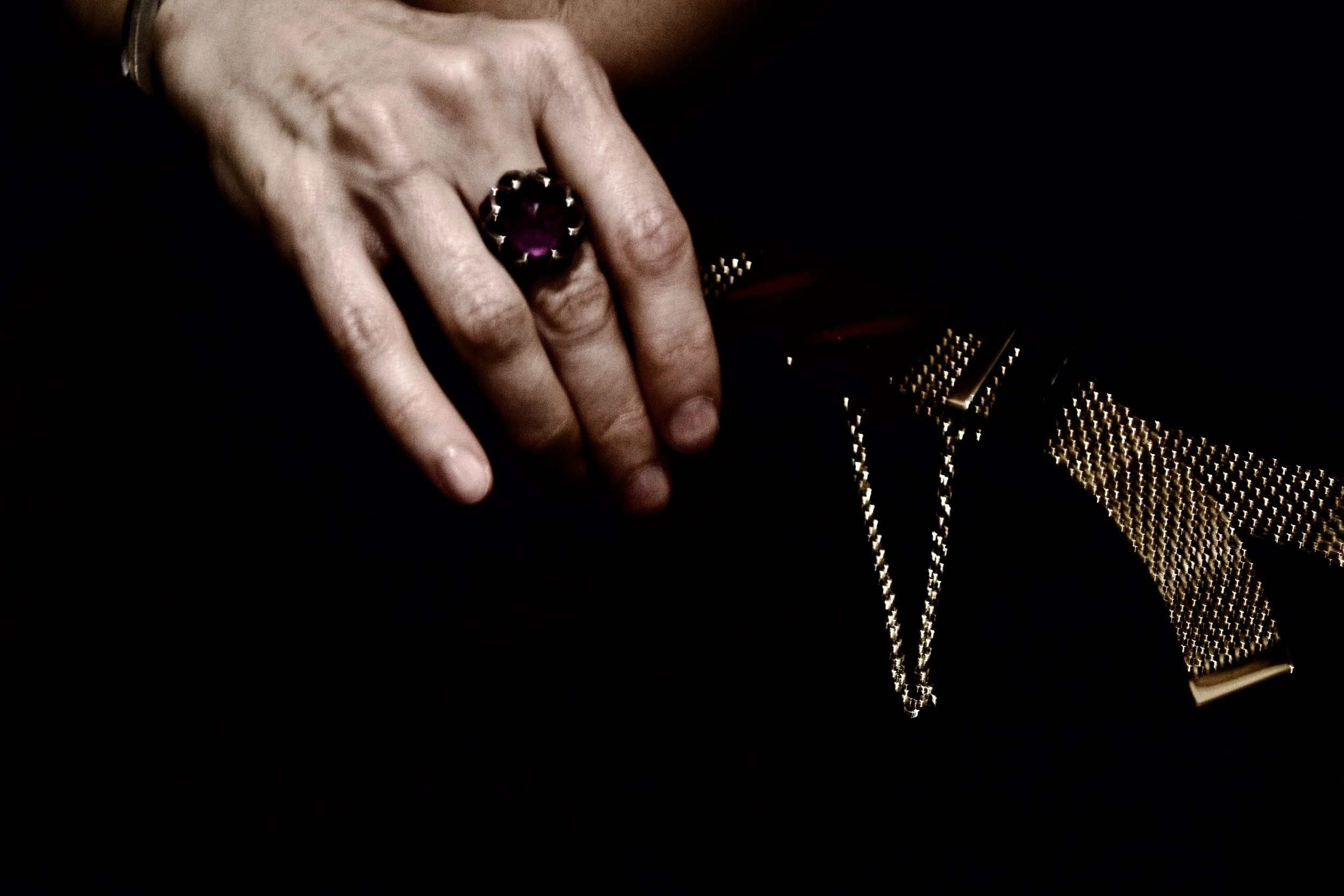Close-up of a woman's hand wearing a large purple gemstone ring, resting on a dark fabric with a gold chain accessory.