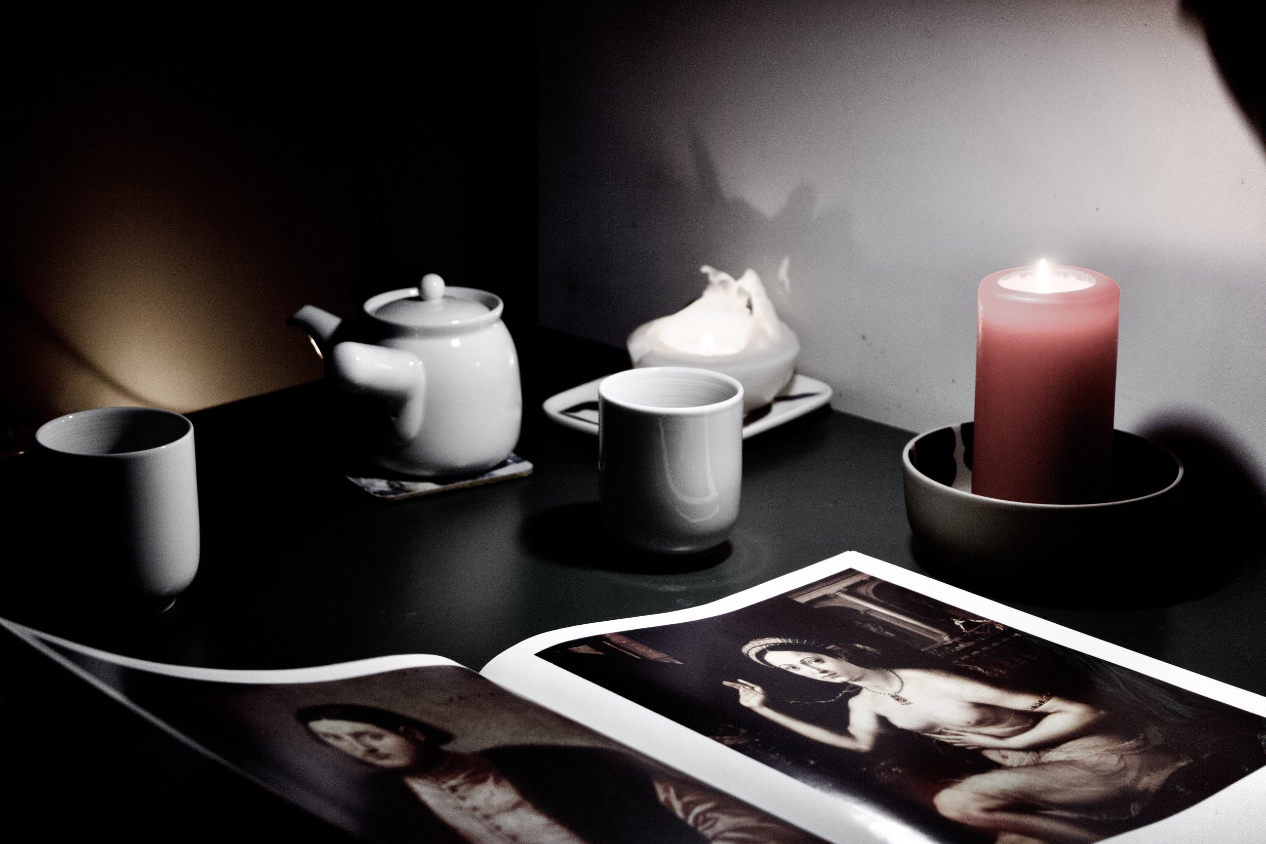 Objects on a table lit by a candle include a teapot, two cups, a small dish, and a magazine with black-and-white photographs.