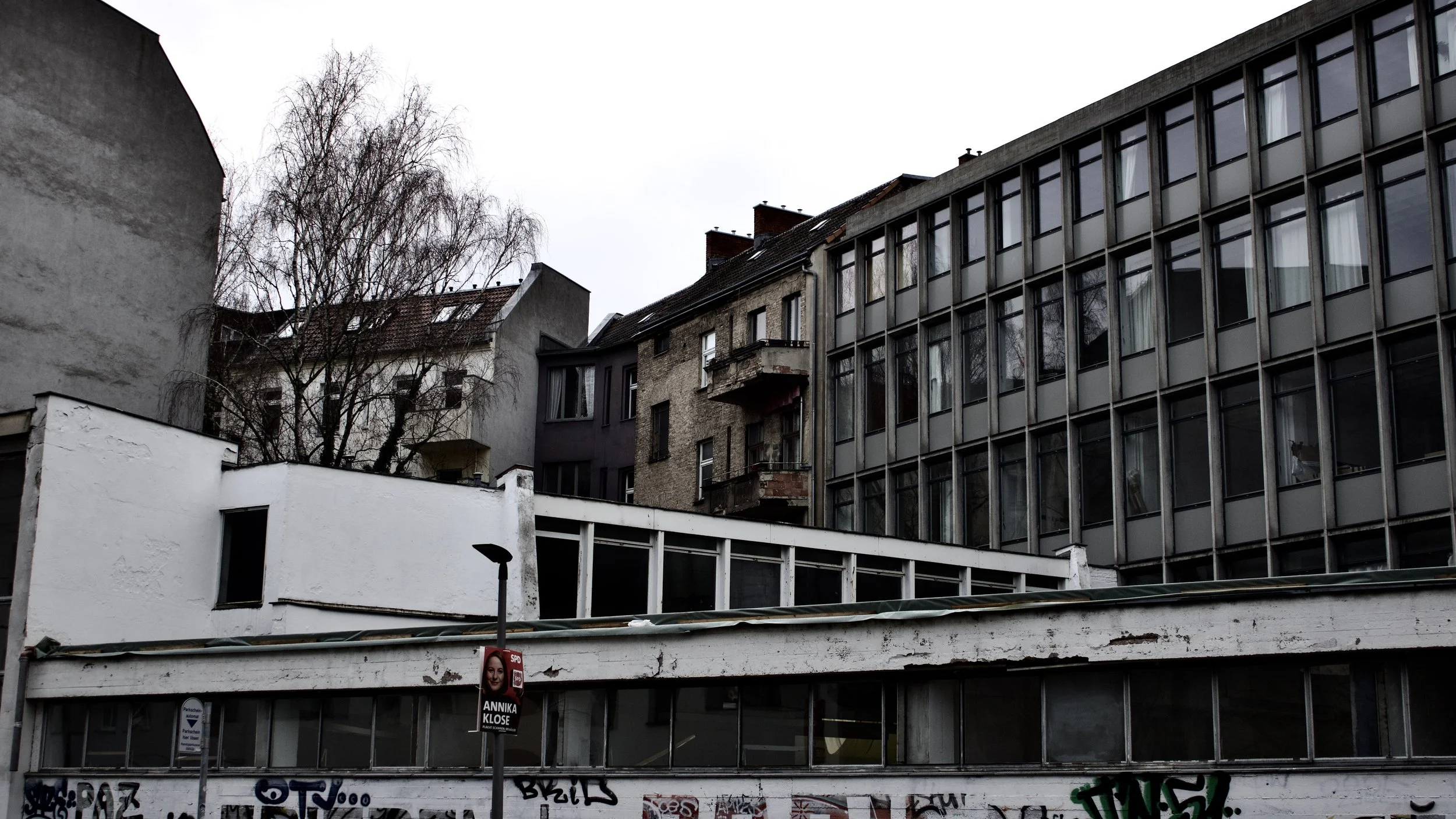 A cityscape featuring a mix of modern and older buildings with multiple windows, a leafless tree against an overcast sky, and street-level graffiti on a white wall. There is a pole with a campaign poster on it in the foreground.
