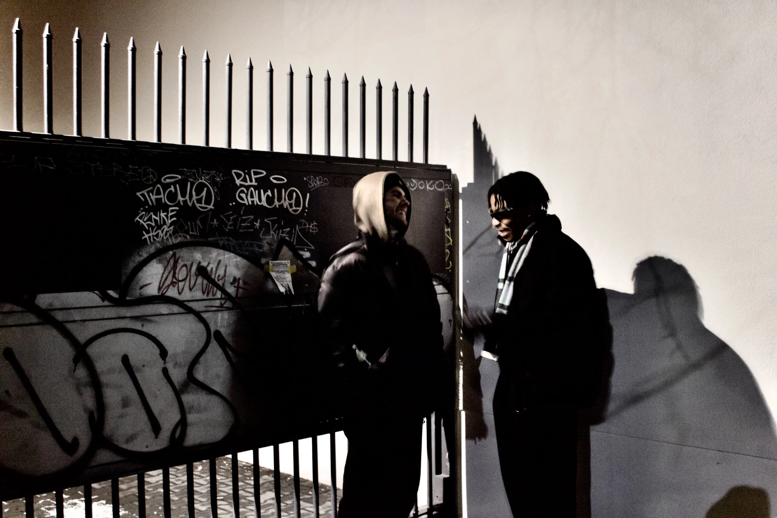 Two people standing near a graffiti-covered fence at night, with tall spikes on top of the fence and shadows cast on a wall behind them.