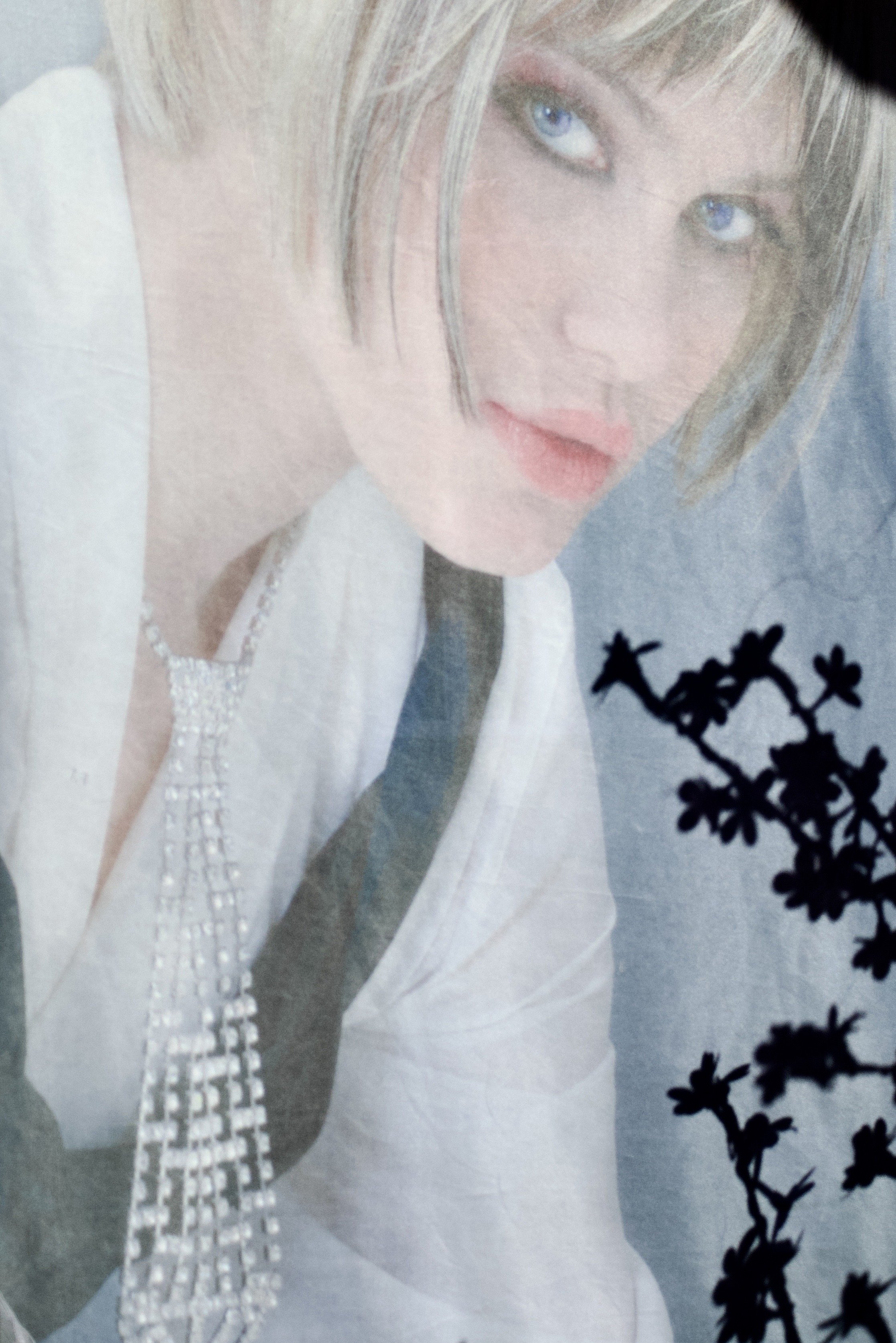 Close-up of a person with blue eyes and blonde hair, wearing a white outfit with pearl embellishments and holding a branch of black flowers.