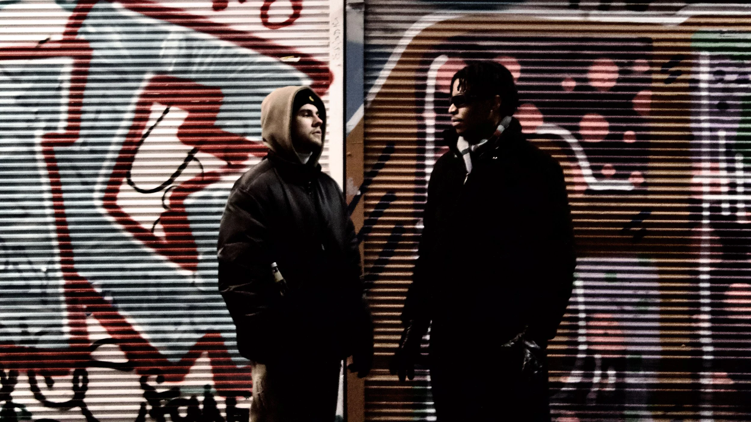 Two young men standing and talking in front of a graffiti-covered metal shutter in a city at night.