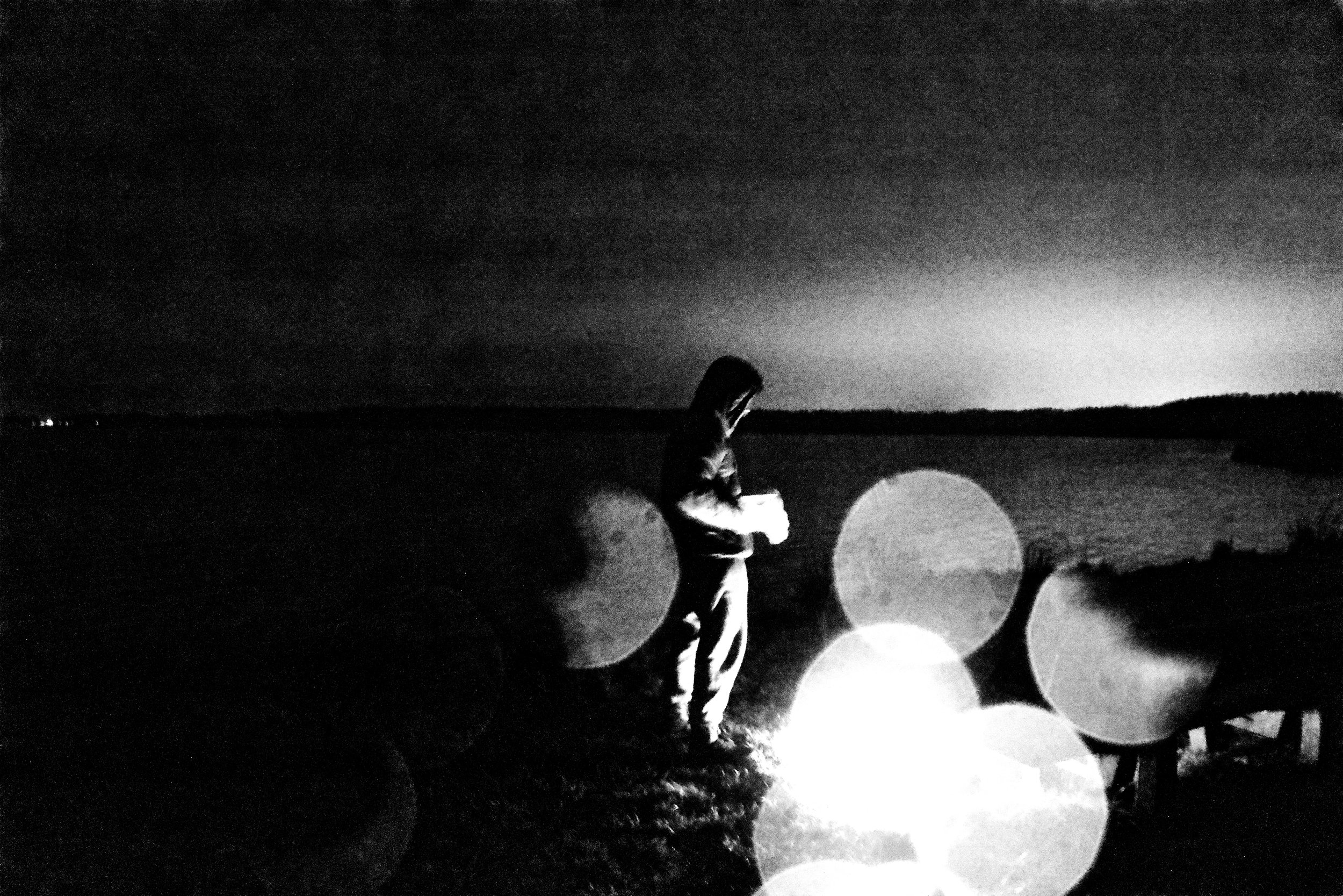 A black and white photo of a person standing outdoors at dusk or night, looking at their phone. There are balloons on the right side of the image, and the background shows a wide open landscape with a horizon. The sky is dark and cloudy.