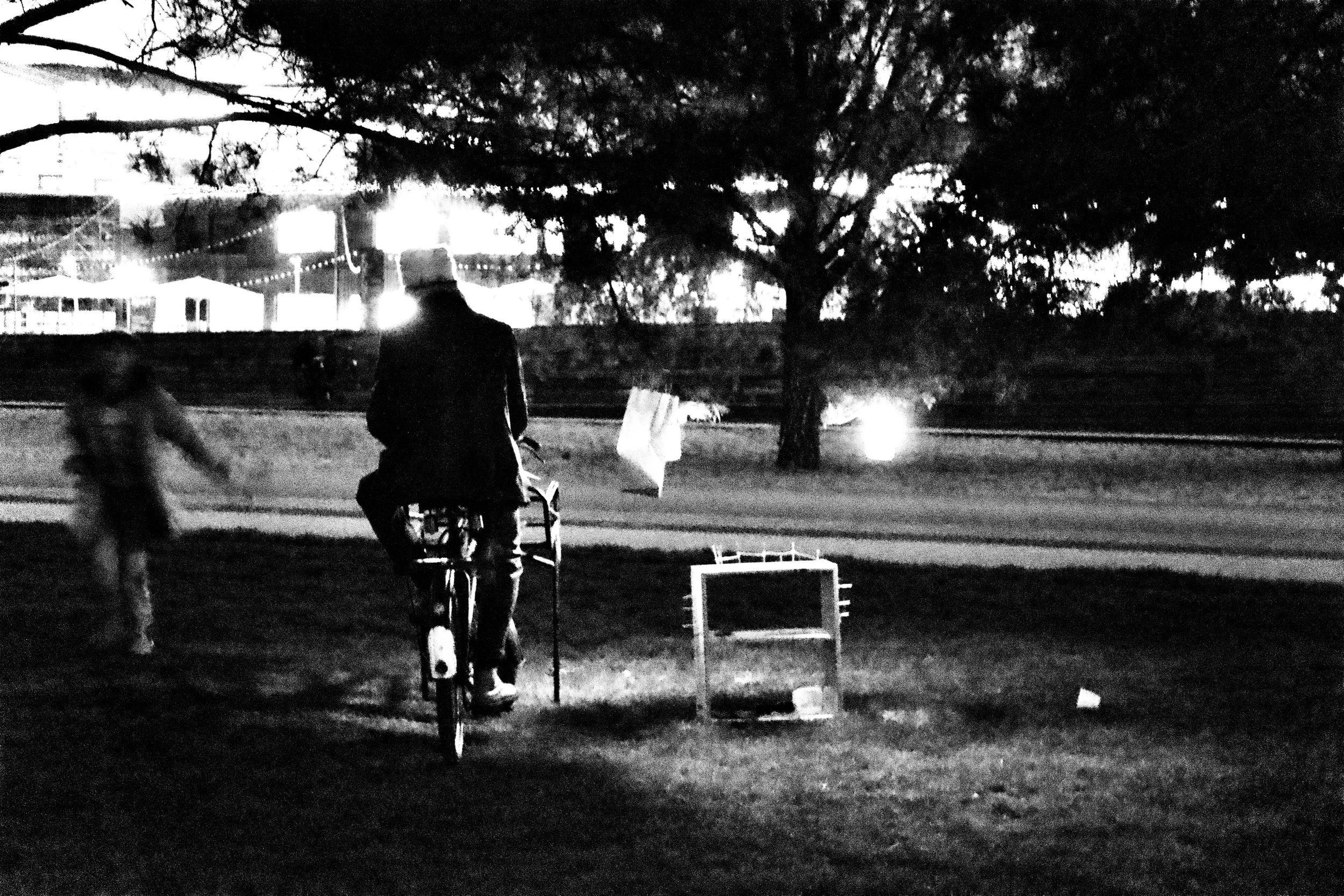 A black and white photo of a person riding a bicycle past an outdoor scene with trees and buildings in the background at night.