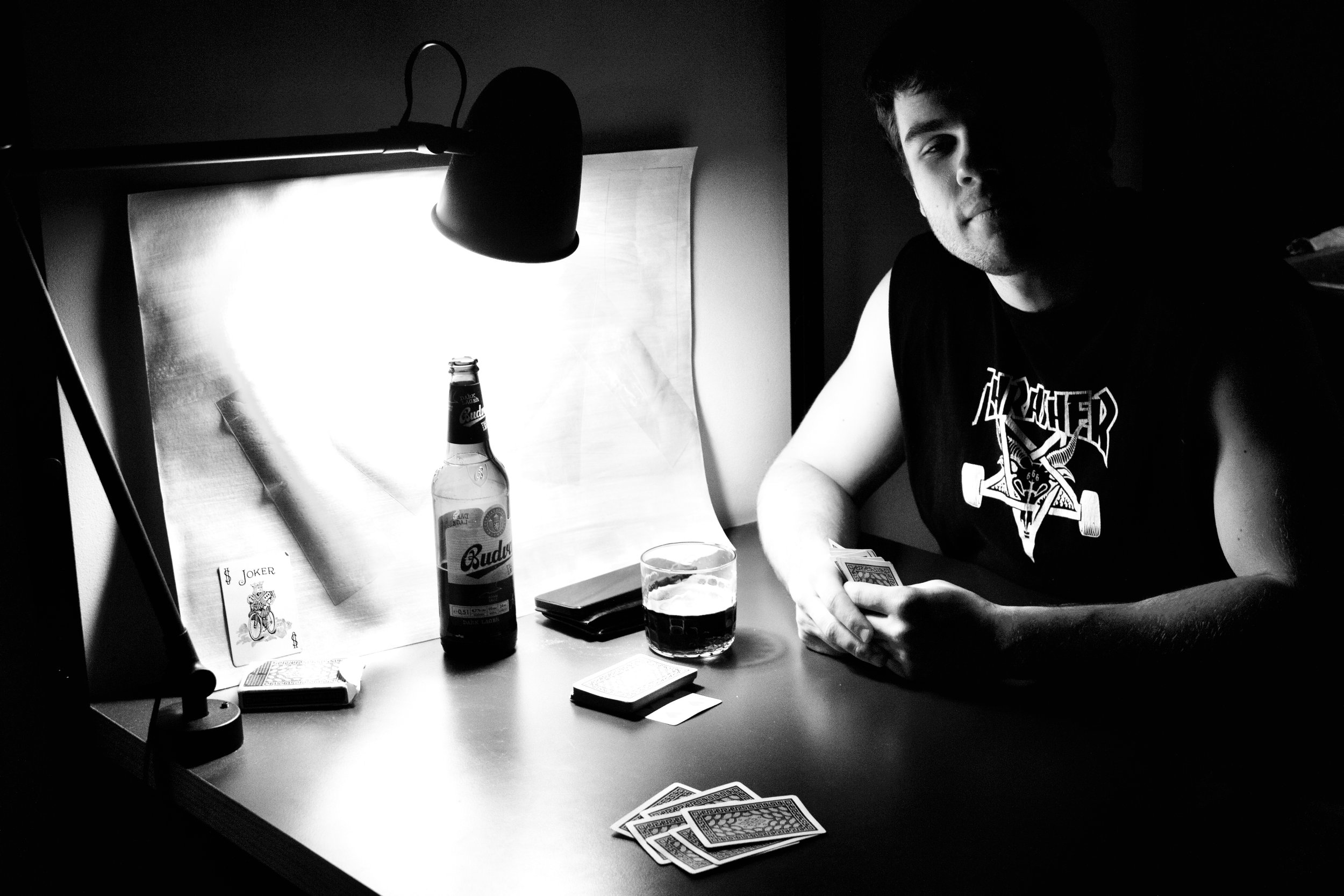 Black and white photo of a young man sitting at a table playing cards, with a beer bottle and a glass on the table. The man is wearing a Thrasher t-shirt and is holding a hand of cards while looking at the camera.