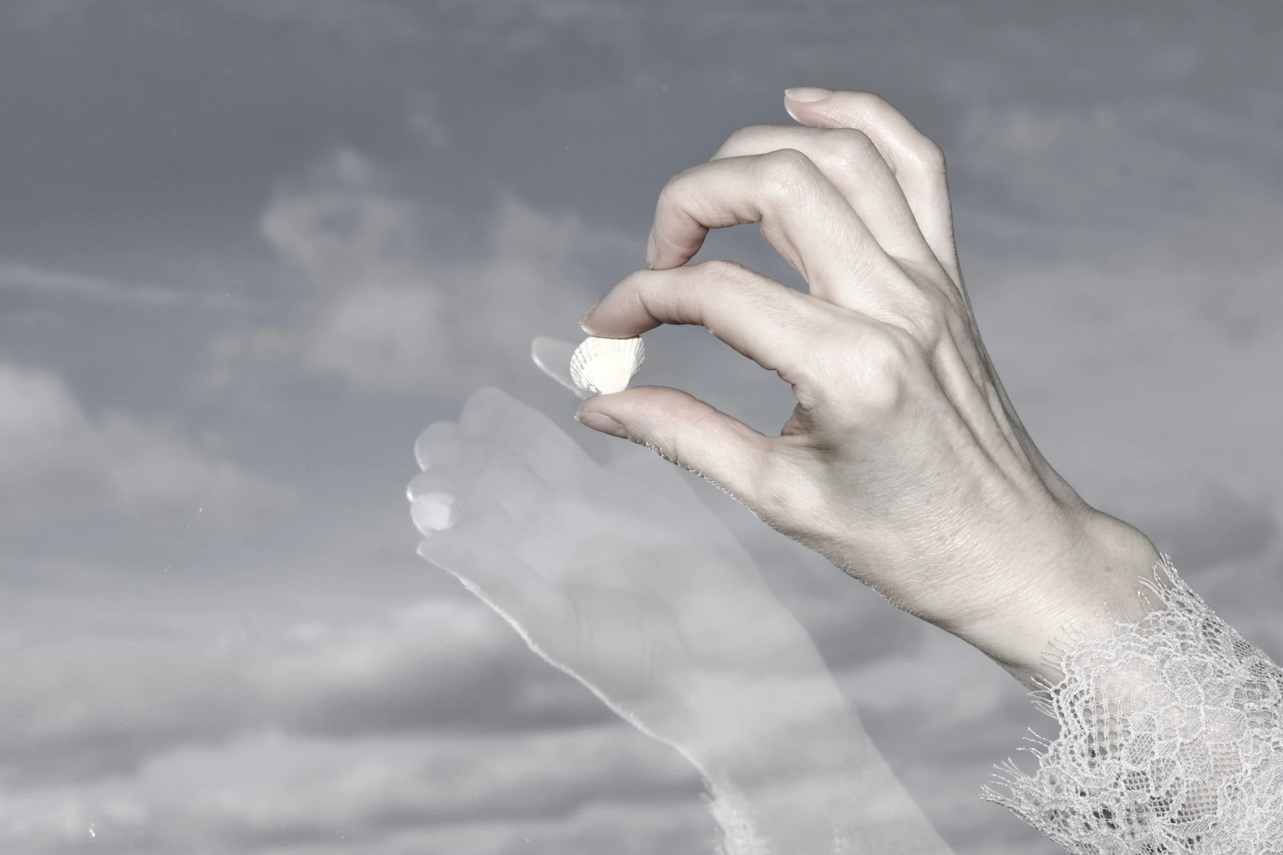 A hand holds a small seashell against a cloudy sky background.