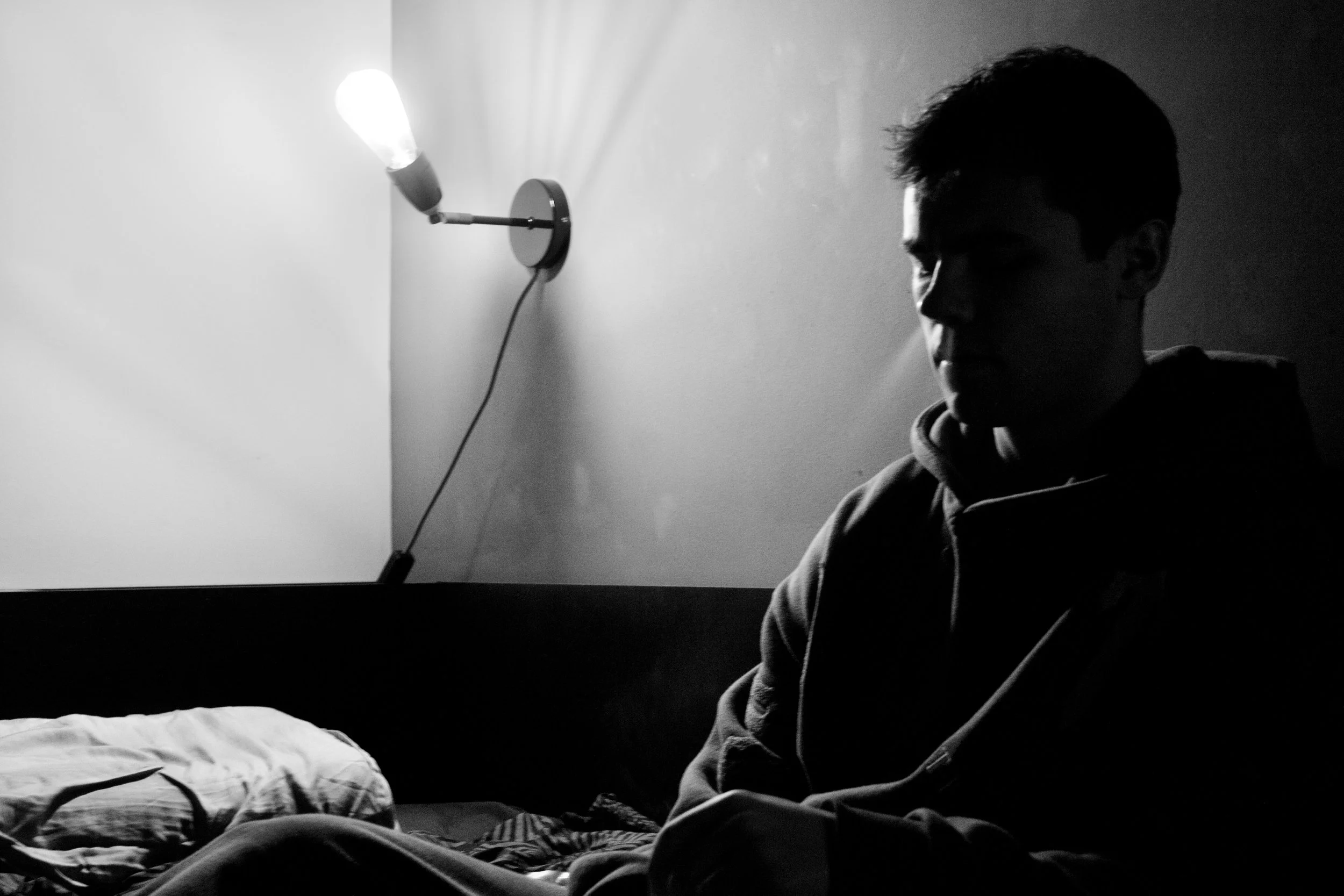 A black and white photo of a person sitting in a dimly lit room, with a wall-mounted lamp on the wall behind them, illuminating parts of the scene. The person appears to be looking down, wearing a hoodie.