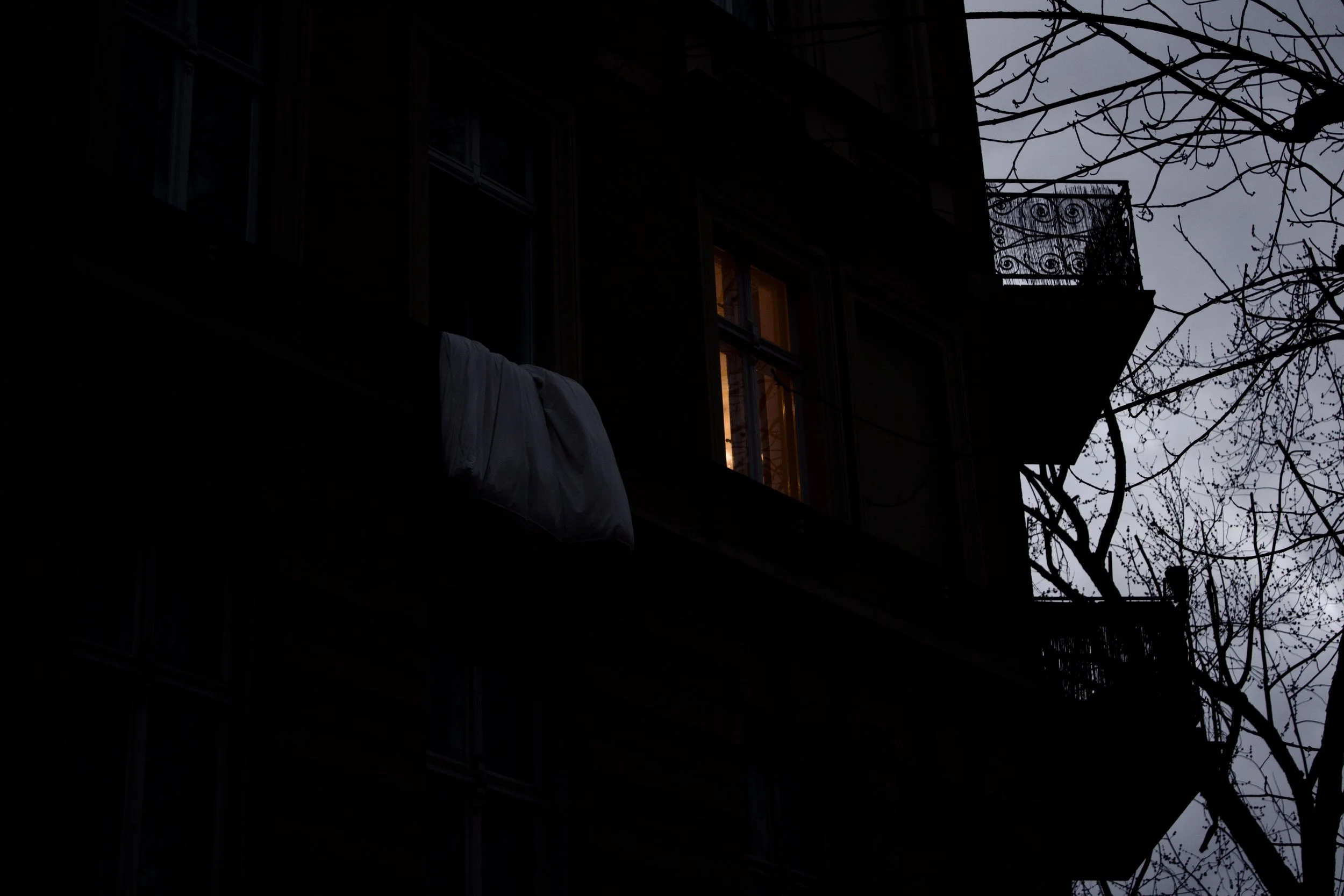 Nighttime view of a multi-story apartment building with a lit window, a balcony with decorative railing, leafless trees, and a cloudy sky.