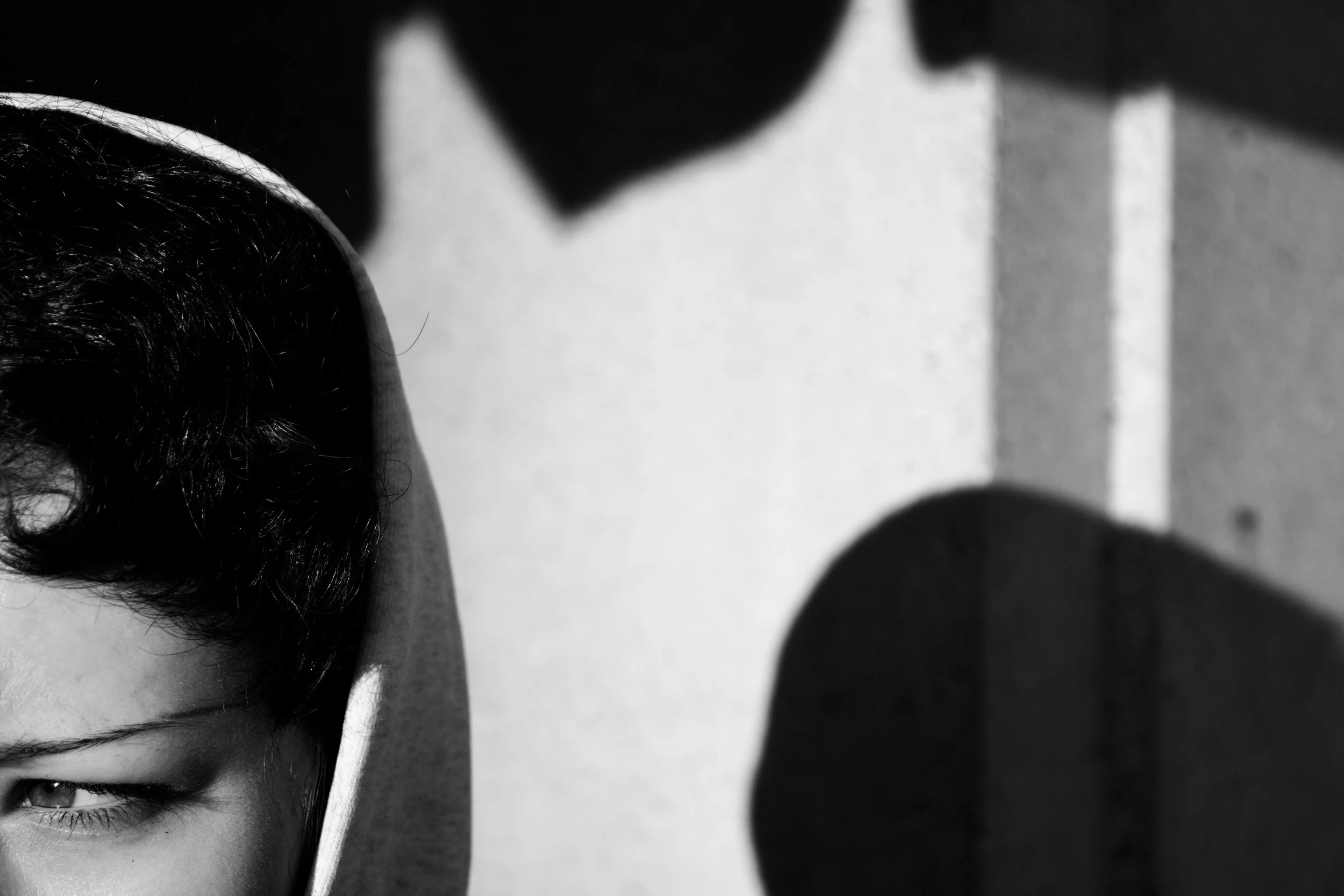 Close-up black and white photo featuring part of a person's face with a focus on the eye and eyebrow in the bottom left corner, and abstract blurred shapes in the background.