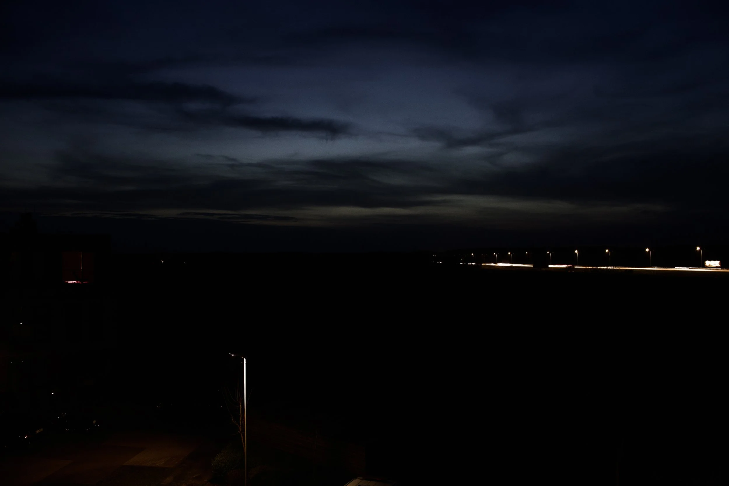 A dark night sky with clouds and city lights near the horizon, with vehicle light trails on a highway.