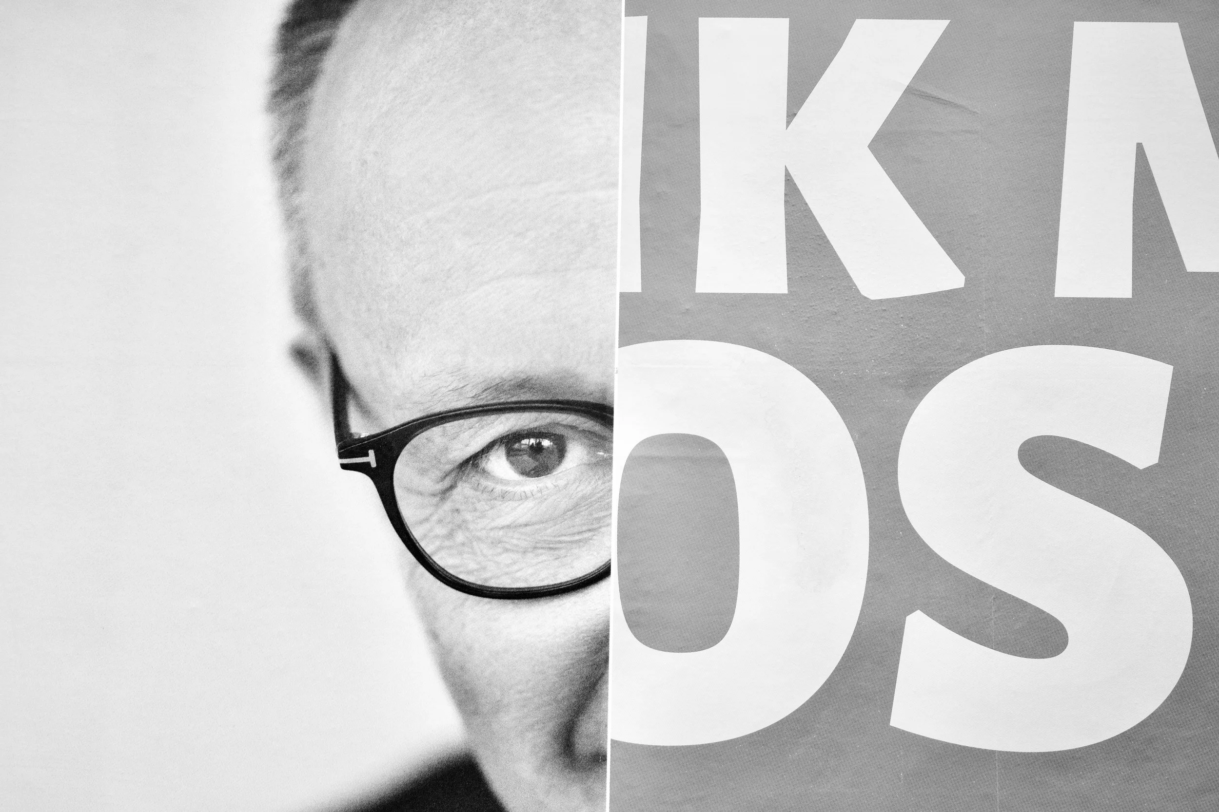 A black and white photo of a person's face, showing only the right eye and part of the face, with large glasses. Overlaying the image are large, bold, white letters spelling out words.