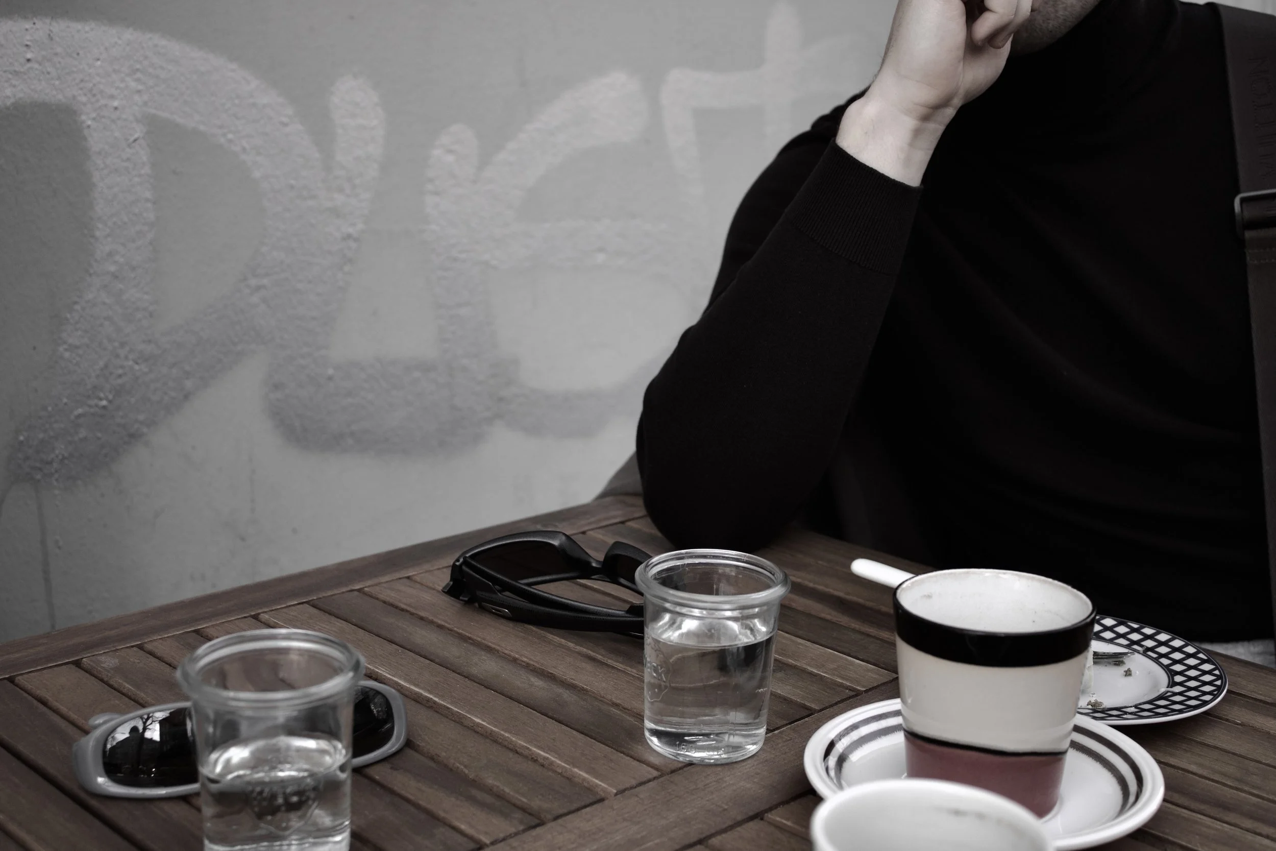 Person sitting at a wooden table with glasses of water, a spoon, and a striped mug, resting their chin on their hand in a black shirt.