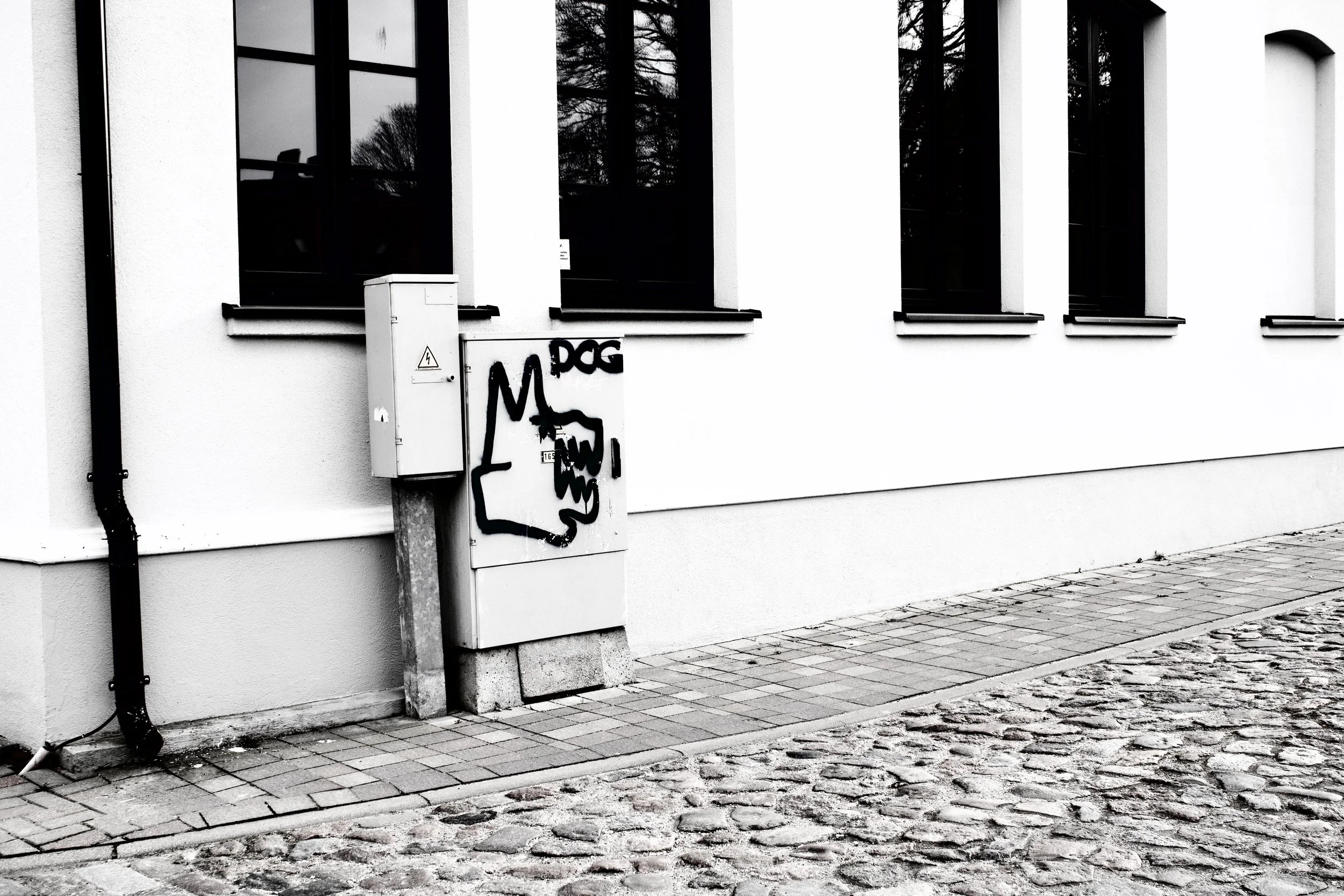 Black and white photograph of a modern building's exterior with four narrow windows and graffiti on a utility box in front. The graffiti shows a stylized face and the words 'M DOG' on the box. A sidewalk with cobblestone and paving stones runs along 