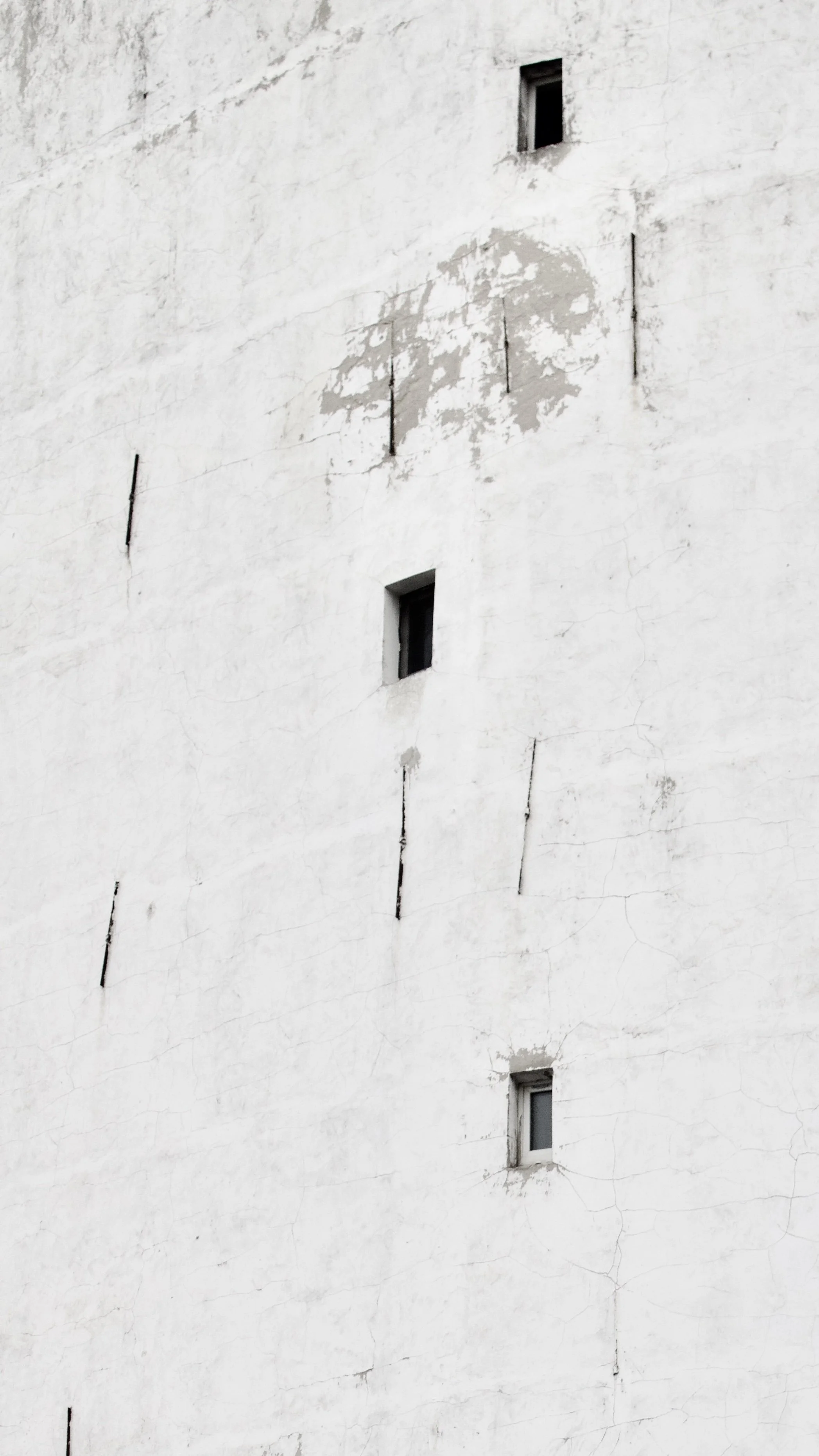 Close-up of a white, weathered building wall with small, varying rectangular and square windows and peeling paint.