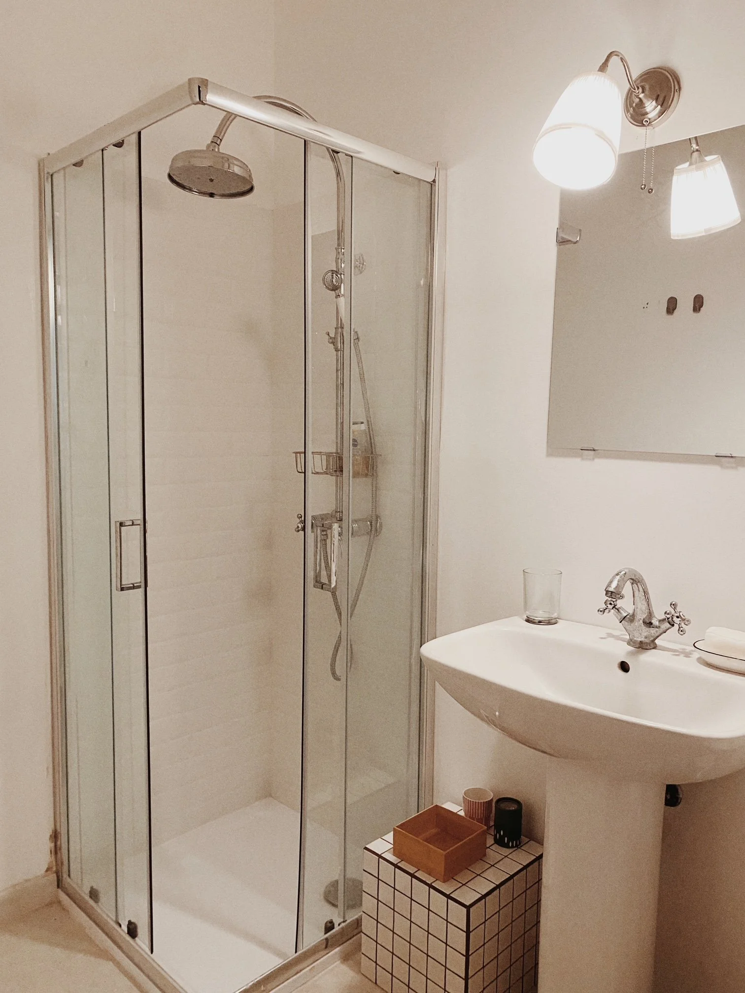 A bathroom featuring a glass-enclosed shower on the left, a white pedestal sink with a faucet, a glass on the sink, and a small tiled side table with a wooden box and two candles. There is a mirror with two light fixtures above it on the wall to the 