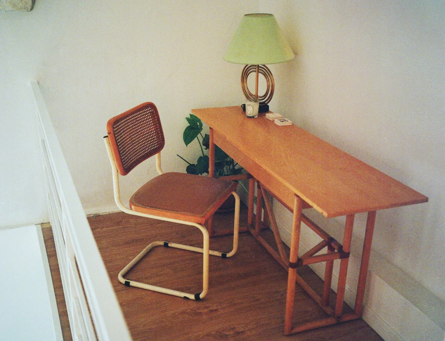 Our cosy working space is made of a wooden desk with a green lamp, a clock, and some small objects, next to a brown chair with a cane backrest, on a wooden floor against a plain white wall.