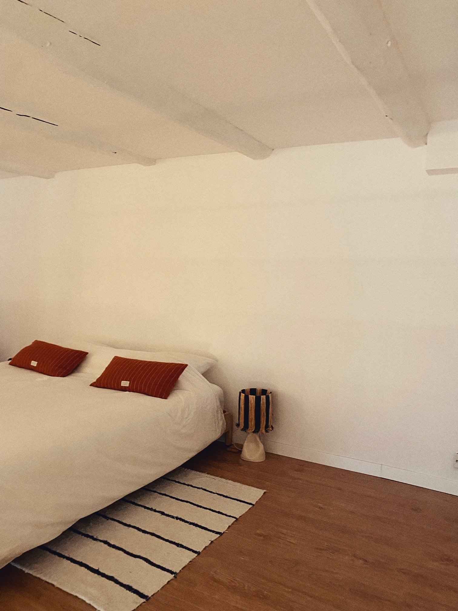 A bedroom with a bed, two reddish-brown pillows, a small striped lamp on a stool, a striped rug, and a white wall with a textured ceiling.