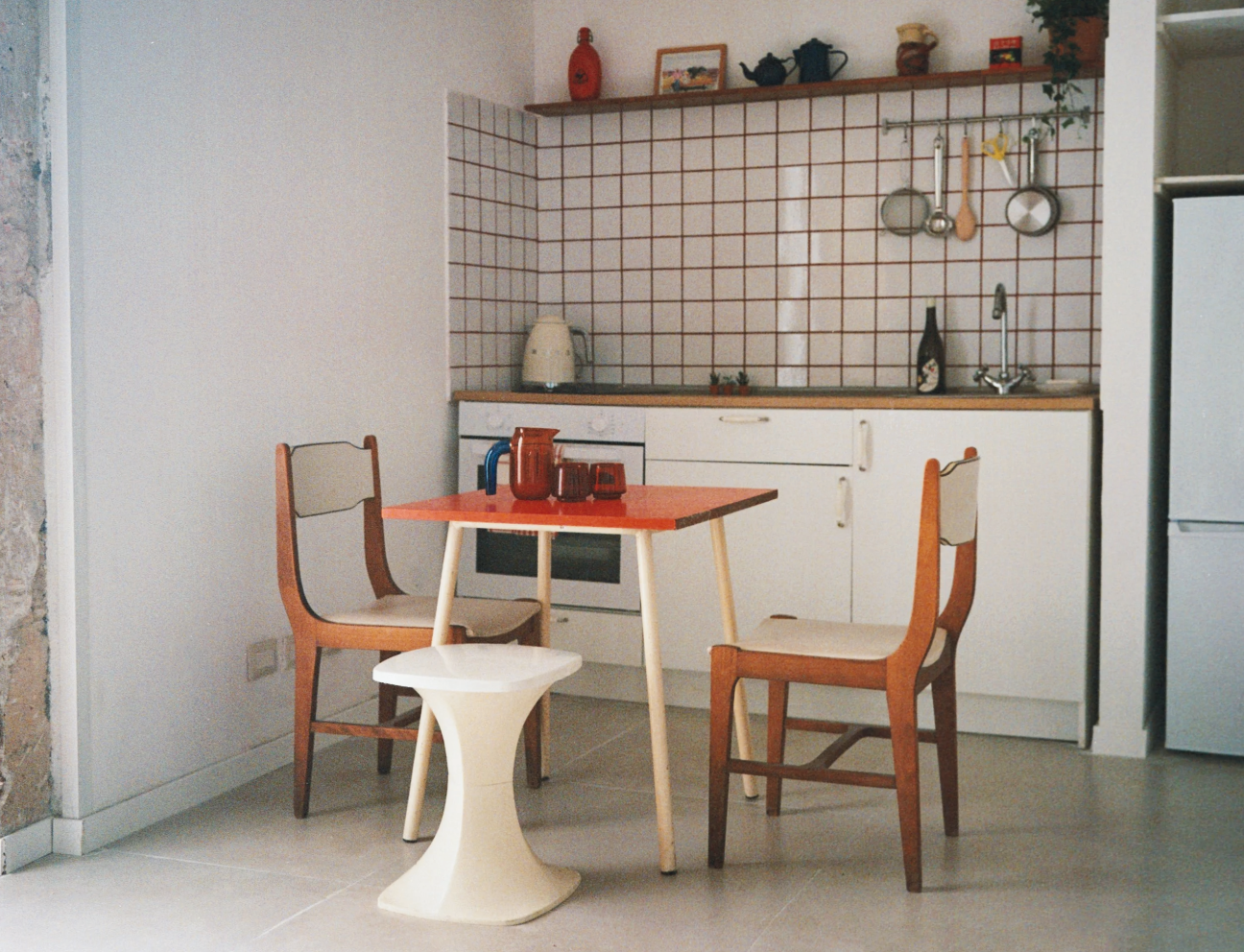 The open kitchen is made with wooden table, two chairs, a white cabinet, and a stove. The backsplash has red and white tiles, with hanging pots and utensils. A pitcher, bottle, and small potted plants are on the countertop.