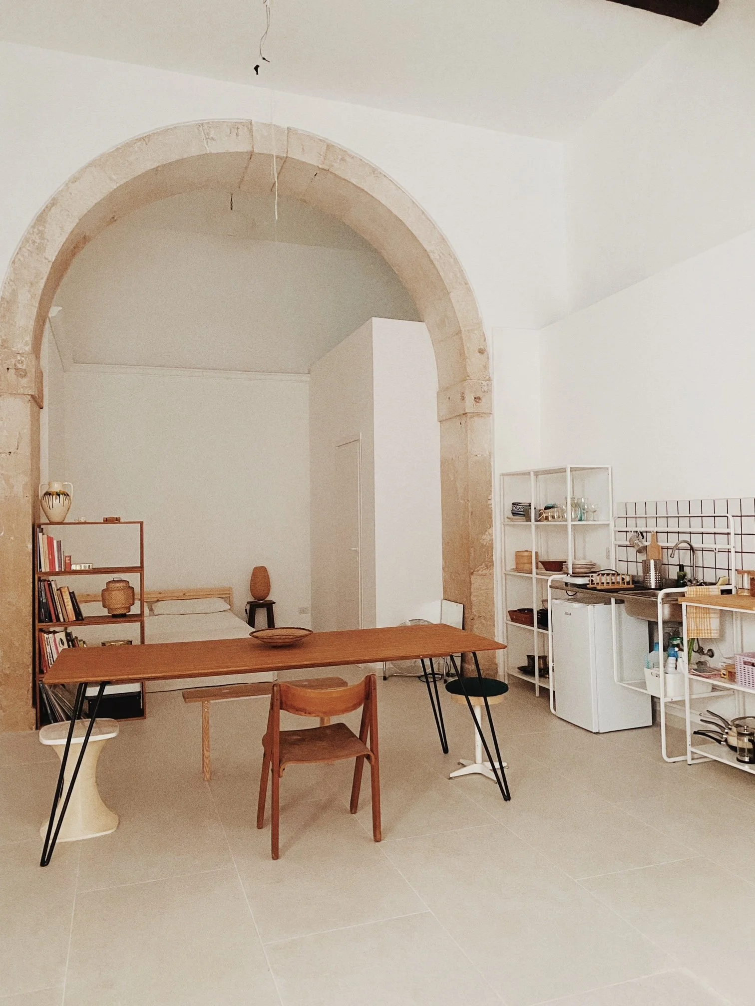 Minimalist kitchen and dining area with a large stone archway, a wooden table, mixed seating, white shelving, and kitchen essentials.