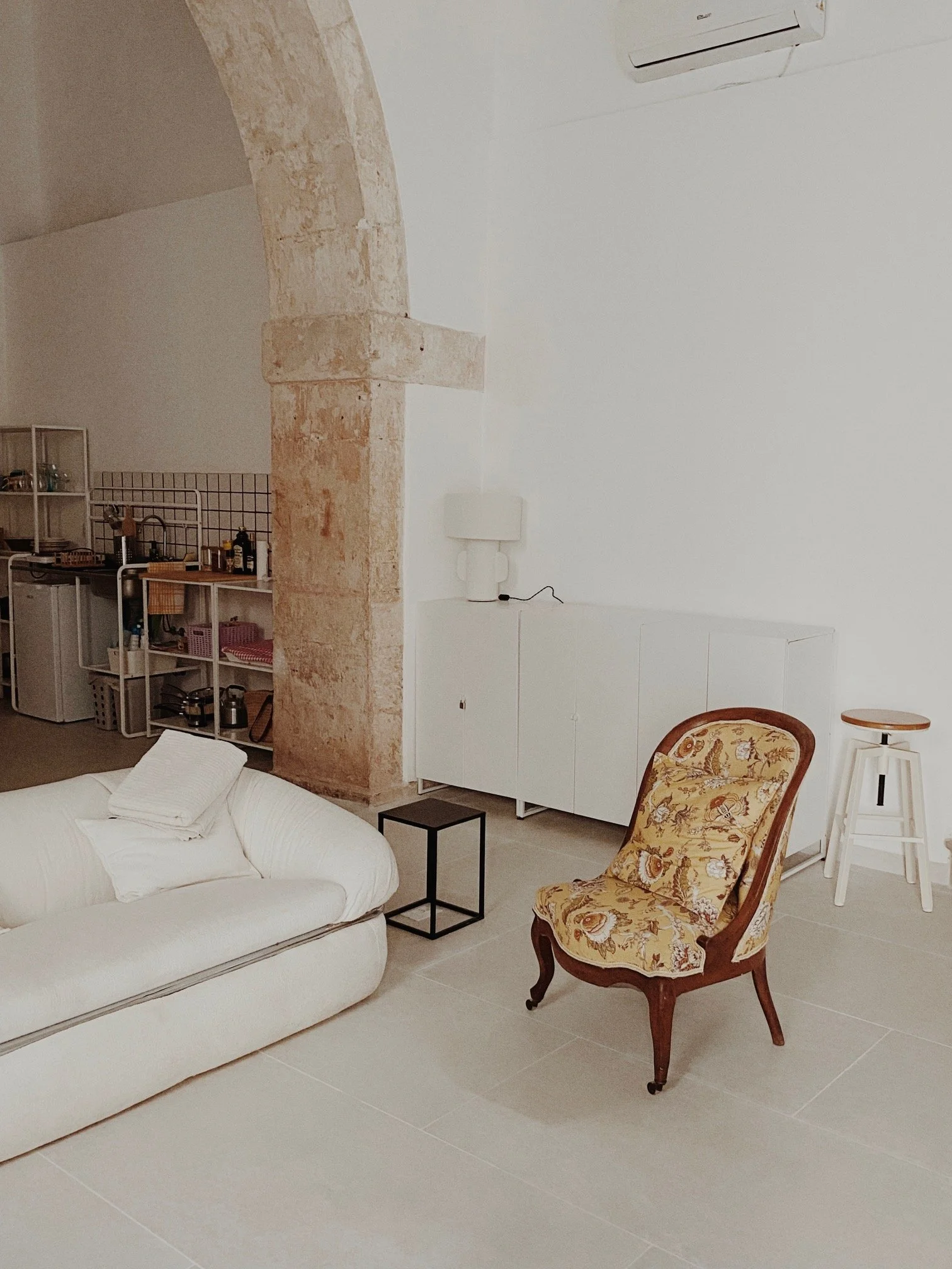 A living room with a white sofa, a patterned armchair, and a small black side table. In the background, there is a kitchen with a sink, shelves, and various kitchen items. The room has a white wall, a stone pillar, and an air conditioning unit on the