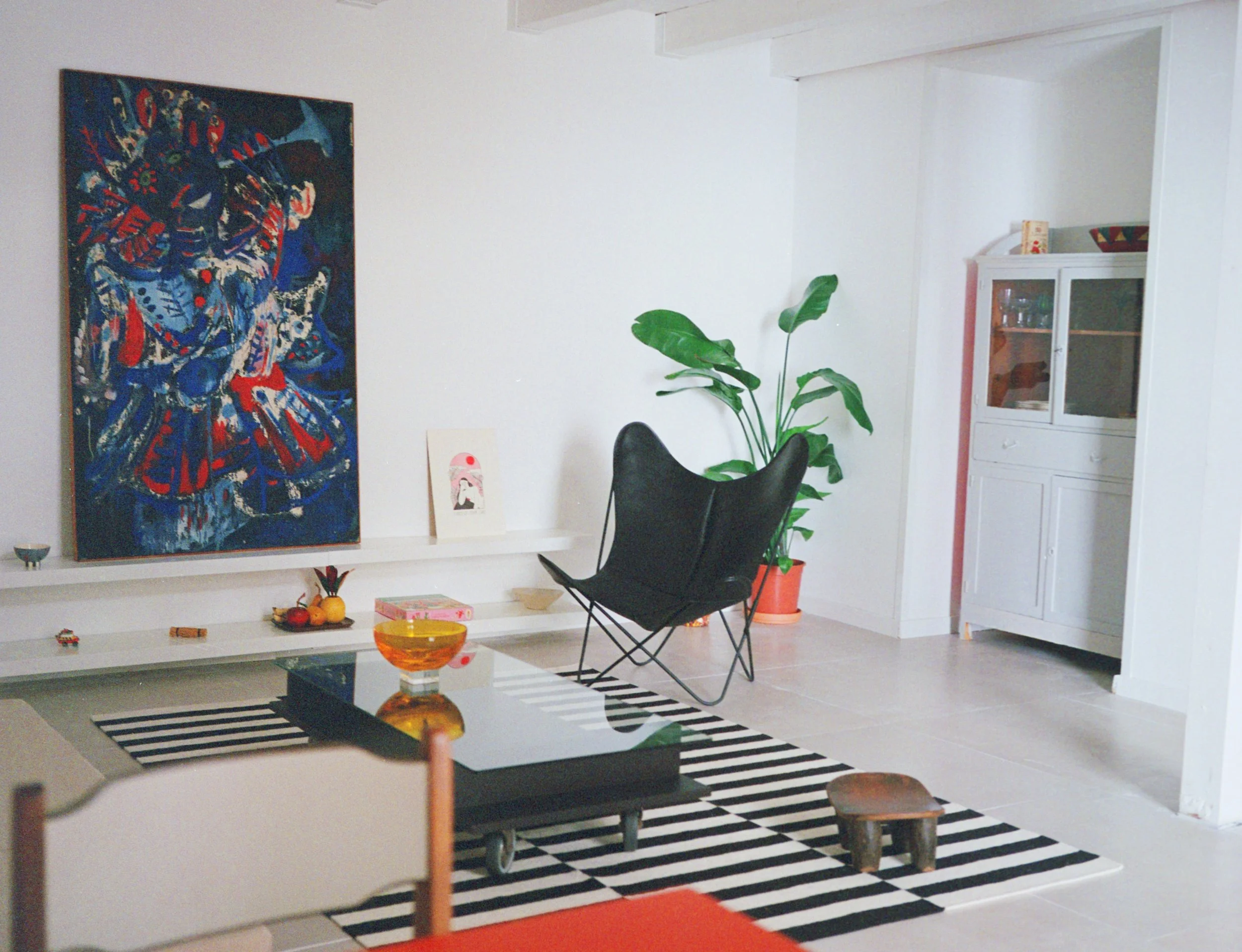Living room with abstract painting, black butterfly chair, green potted plant, striped black and white rug, coffee table with orange bowl, and white cabinet.