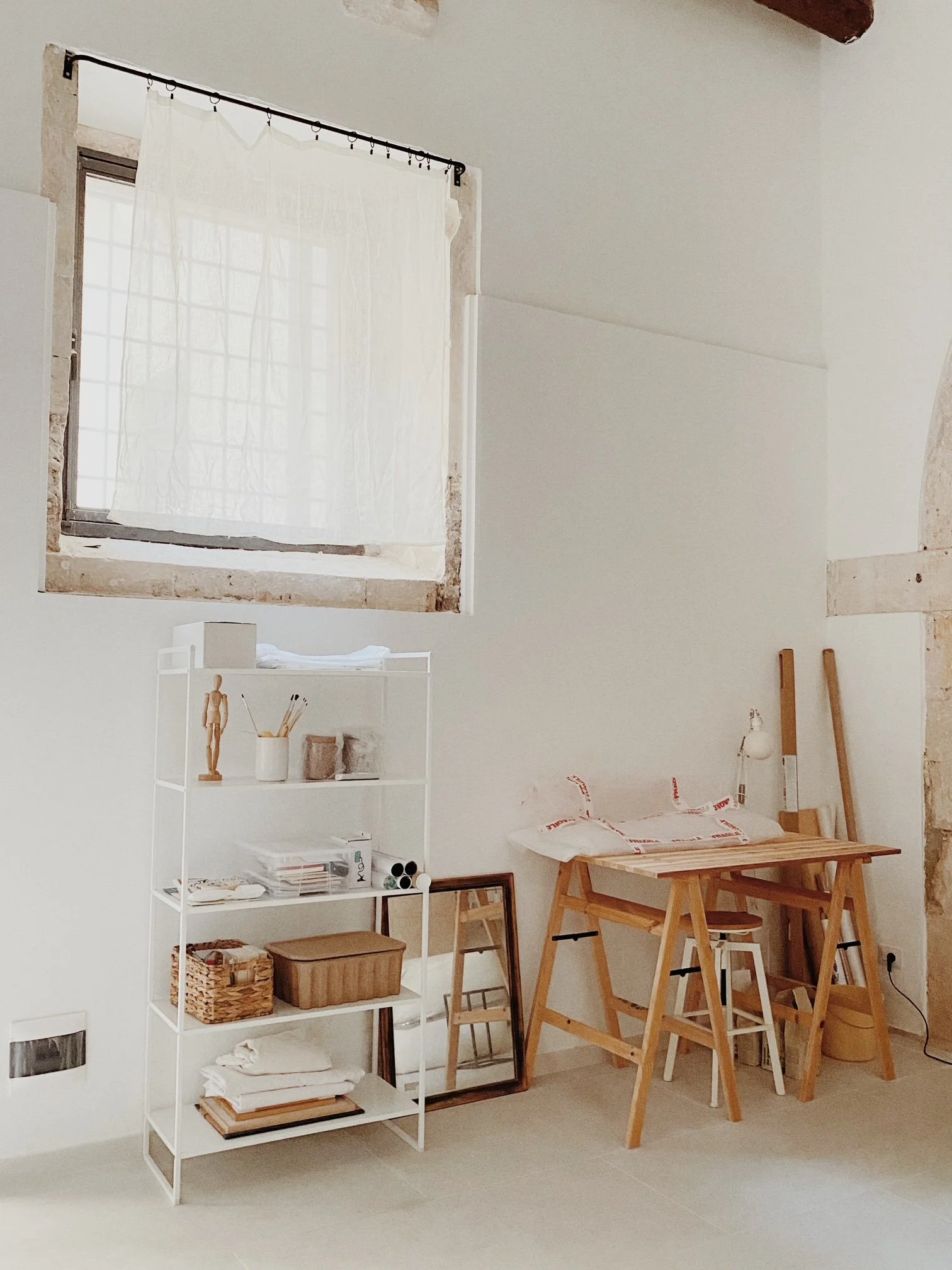 A minimalistic room with a window, white wall, a white shelf with decor and storage boxes, a mirror leaning against the wall, a wooden worktable with a pillow, and some wooden planks and a small stool nearby.