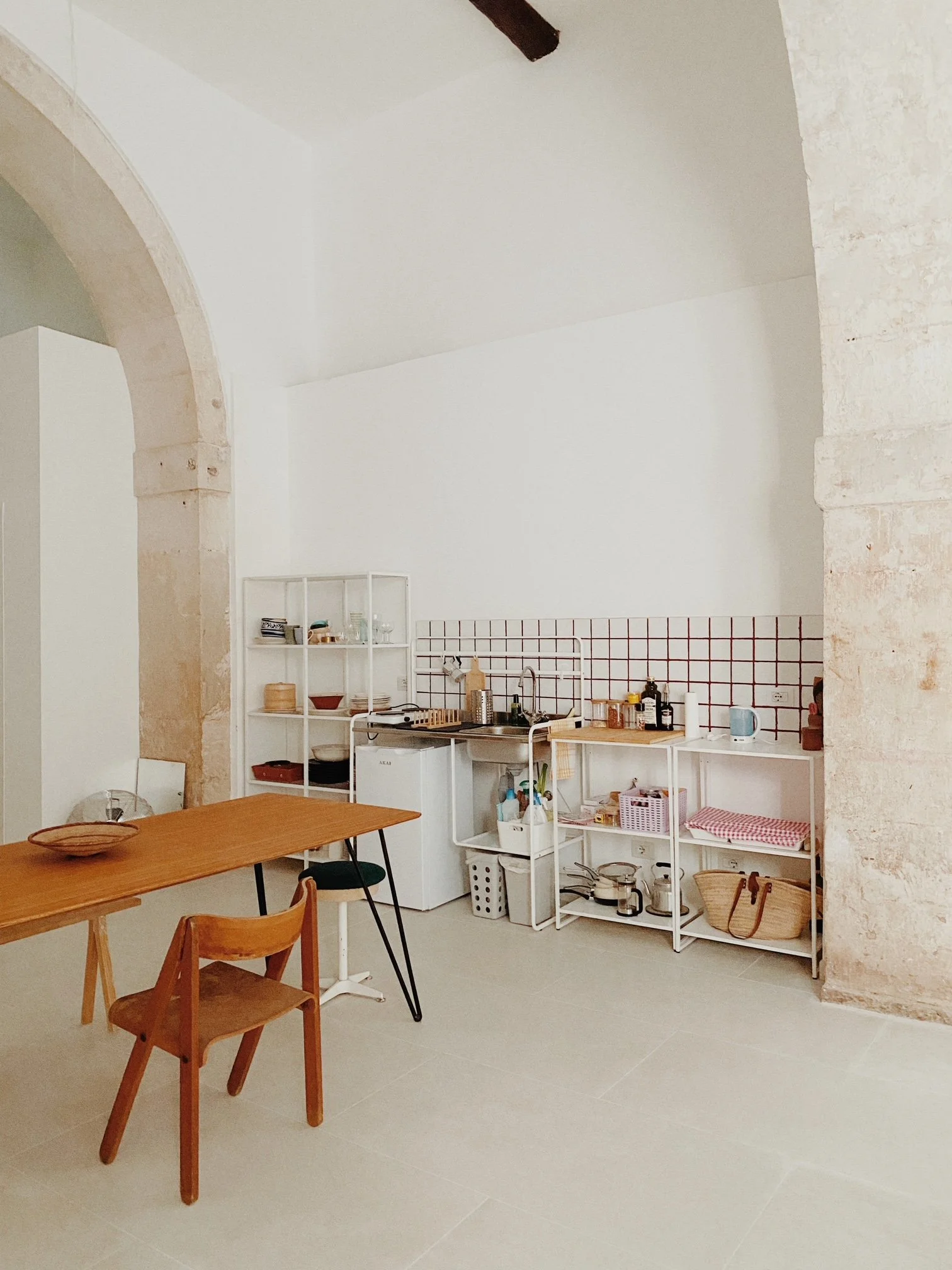 The kitchen area of Lo Studio Alto is made with a small sink, open white shelving, a white mini fridge, and a wooden dining table with two chairs in a room featuring stone arches and white walls.