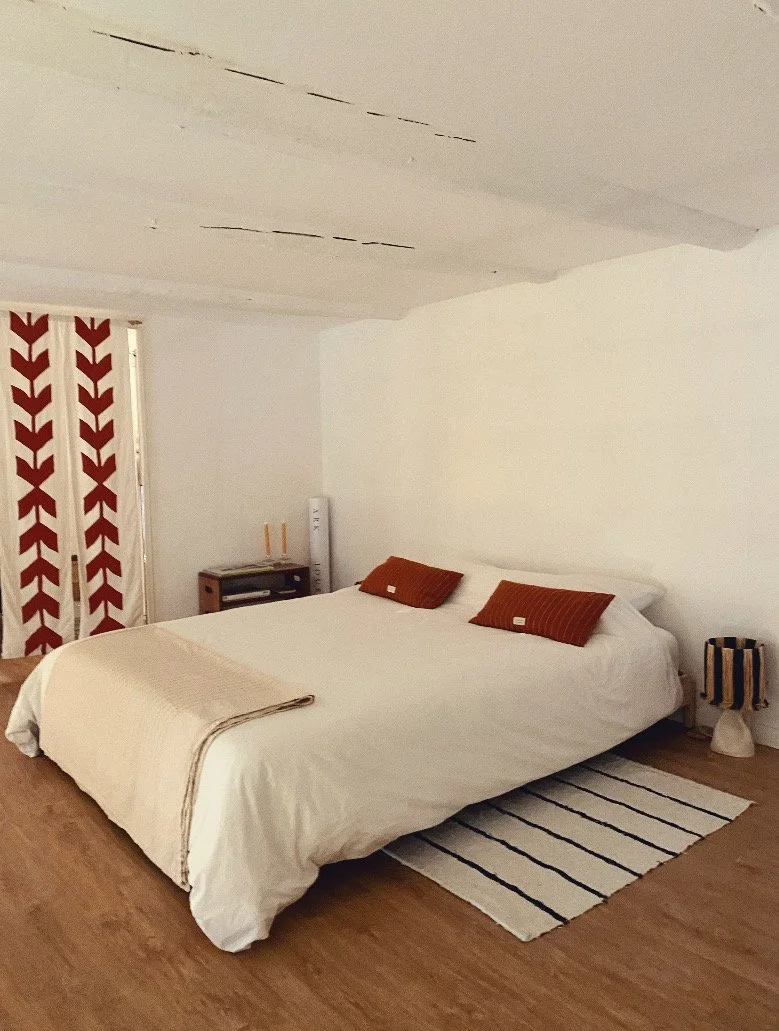 A bedroom with a large bed covered in white bedding, two brown pillows, and a beige throw blanket. There are patterned curtains with red and white geometric designs on a window, a small wooden nightstand with two candles, a tall white heater or fan, 