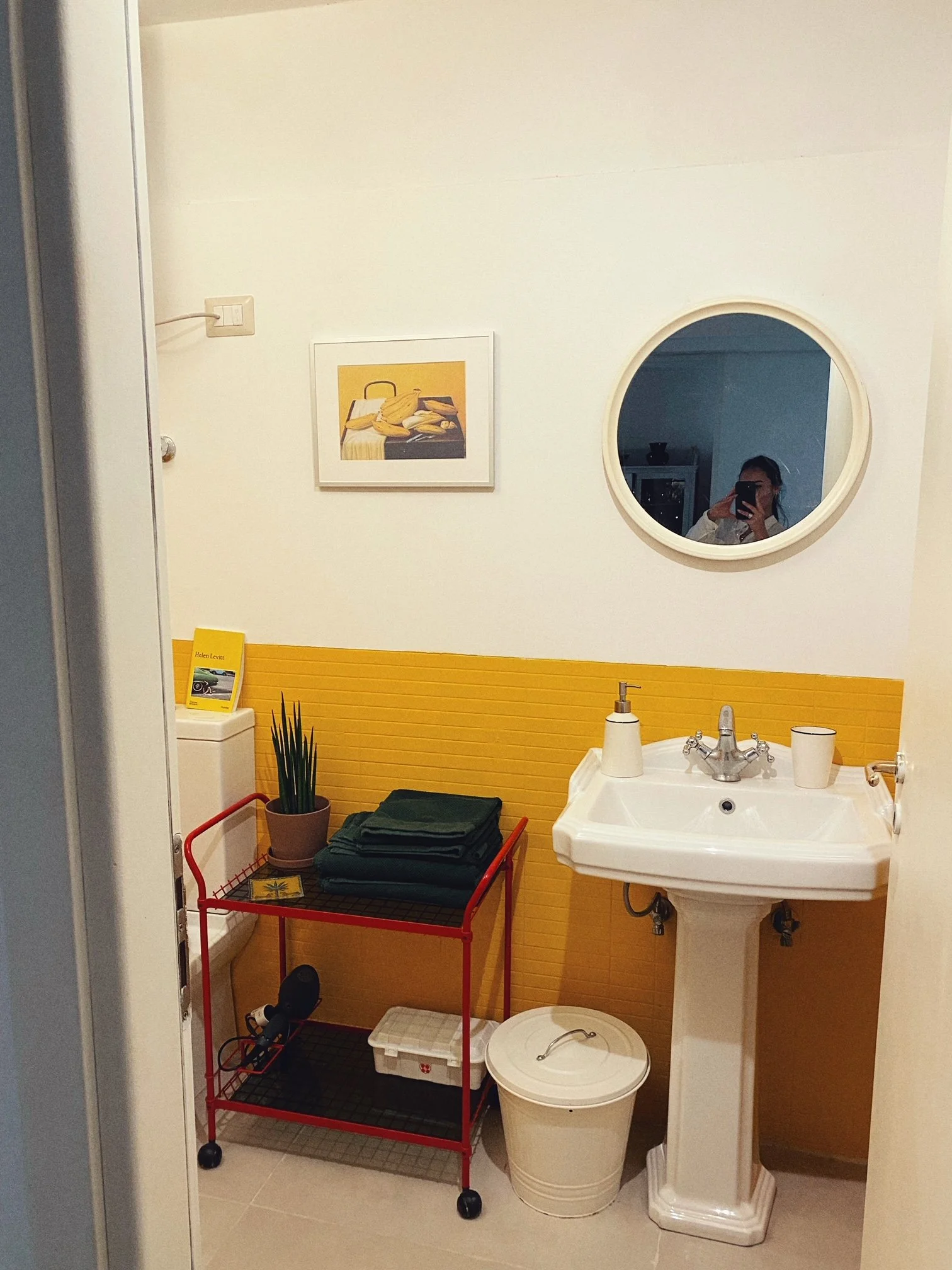A small bathroom with a white pedestal sink, a round mirror, a yellow textured wall panel, a potted plant on a red cart with folded towels, a laundry basket, and a person taking a selfie with a phone in the mirror.