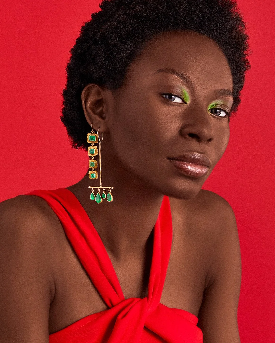 Black woman wearing gold and emerald drop earrings with green eyeshadow against a vibrant red background.