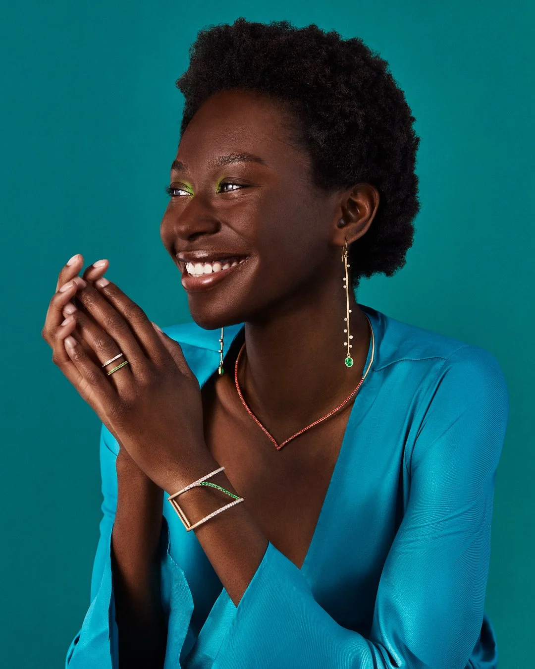 Smiling Black woman wearing elegant drop earrings, a V-shaped necklace, rings, and a bracelet against a teal backdrop