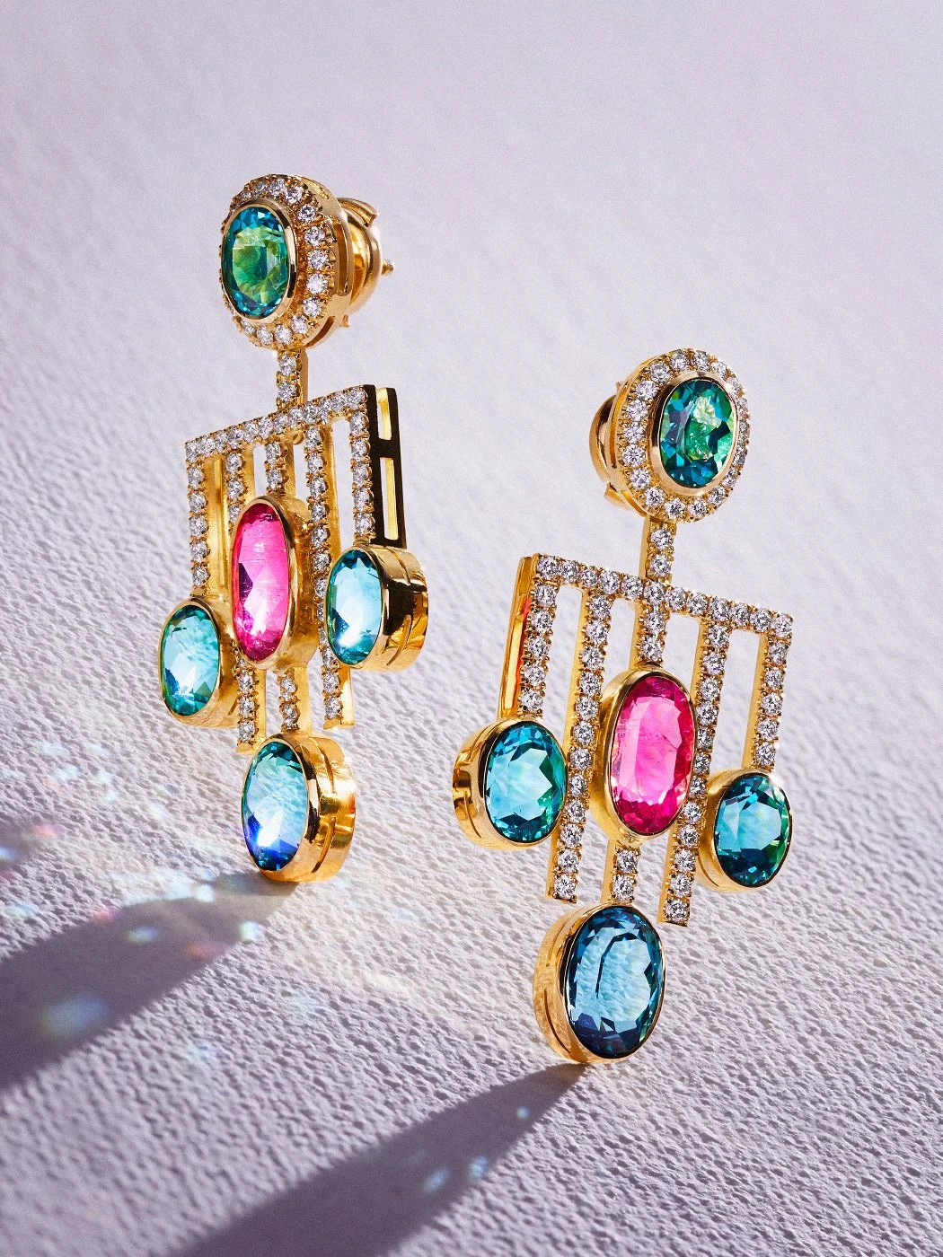 Gold geometric chandelier earrings with pink and blue gemstones and diamond accents on a white background.