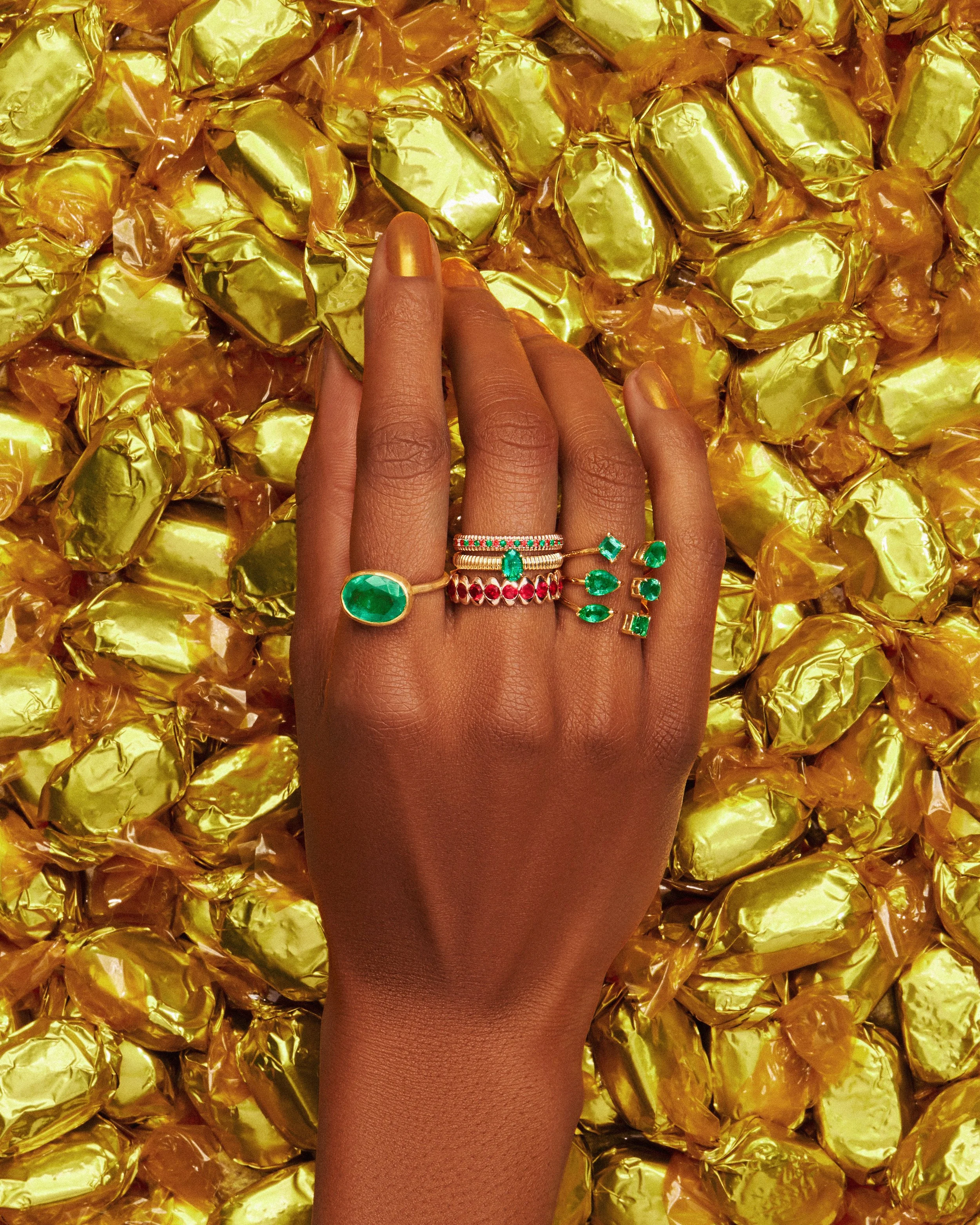 Hand wearing luxury gold rings with emeralds and rubies over a pile of gold-wrapped candies