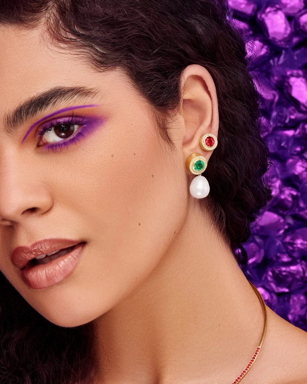 Woman with vibrant purple eye makeup wearing gold earrings with ruby, emerald, and a white pearl drop -  Gemfields campaign. 