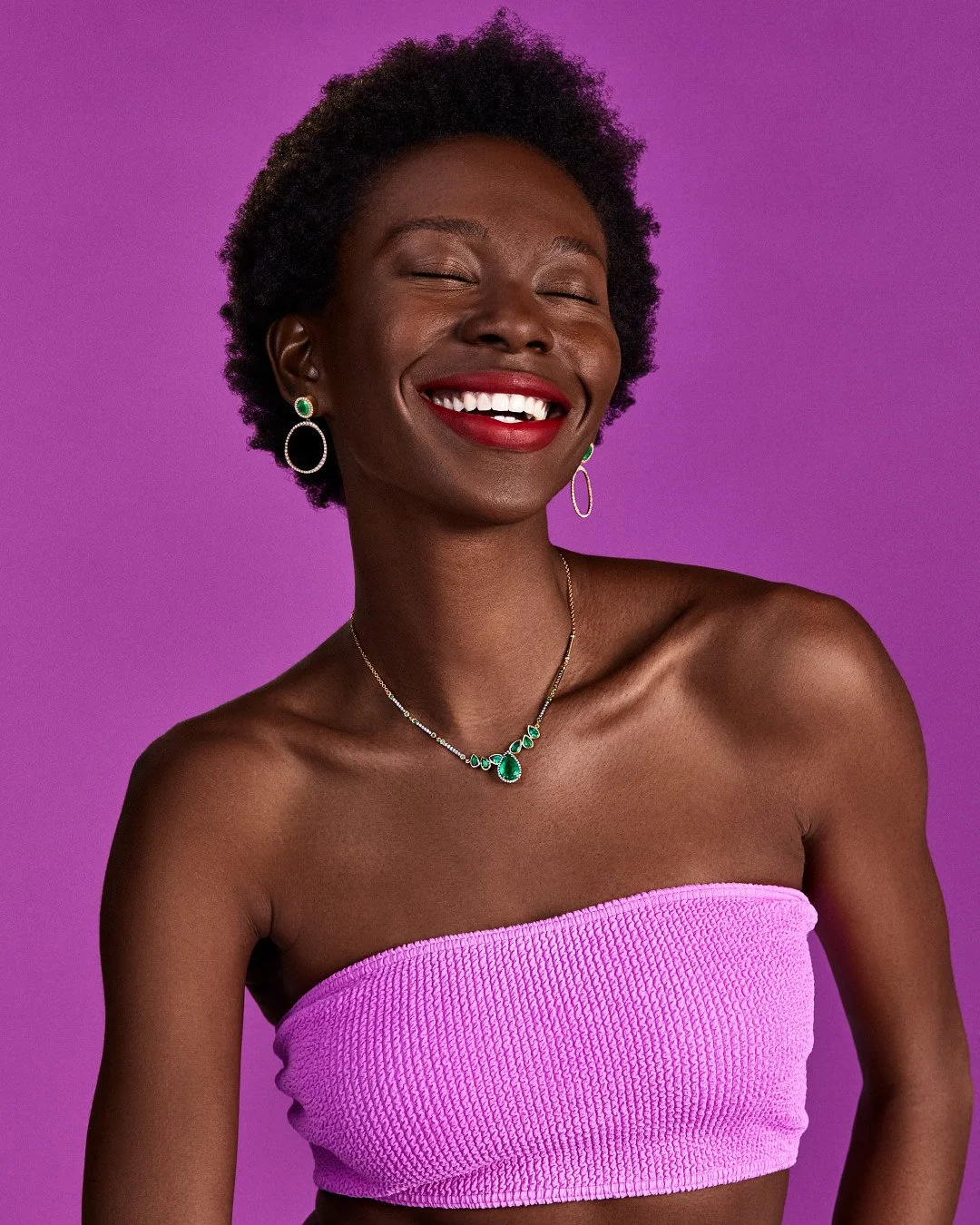 Smiling Black woman wearing an emerald necklace and matching earrings against a vibrant purple background.