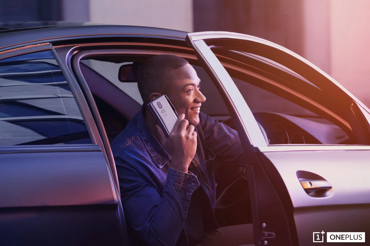 A smiling man sits in the driver's seat of a car while talking on a silver OnePlus smartphone.