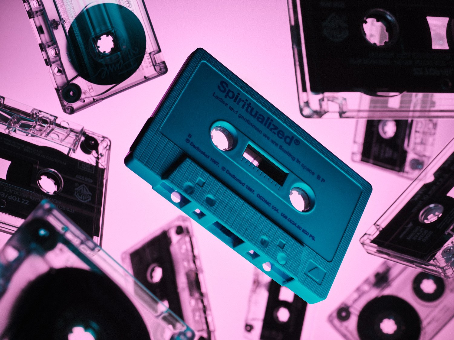 Teal Spiritualized cassette tape floating among other tapes on a neon pink background in retro synthwave style.