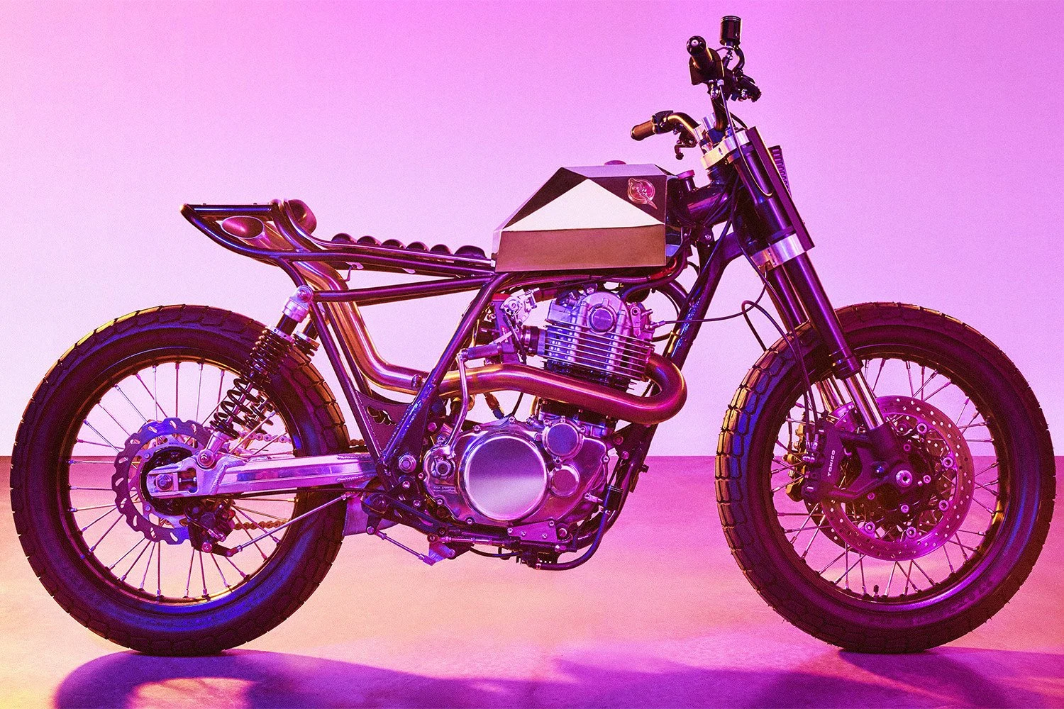 Custom scrambler motorcycle with geometric silver fuel tank under vibrant pink and magenta studio lighting.