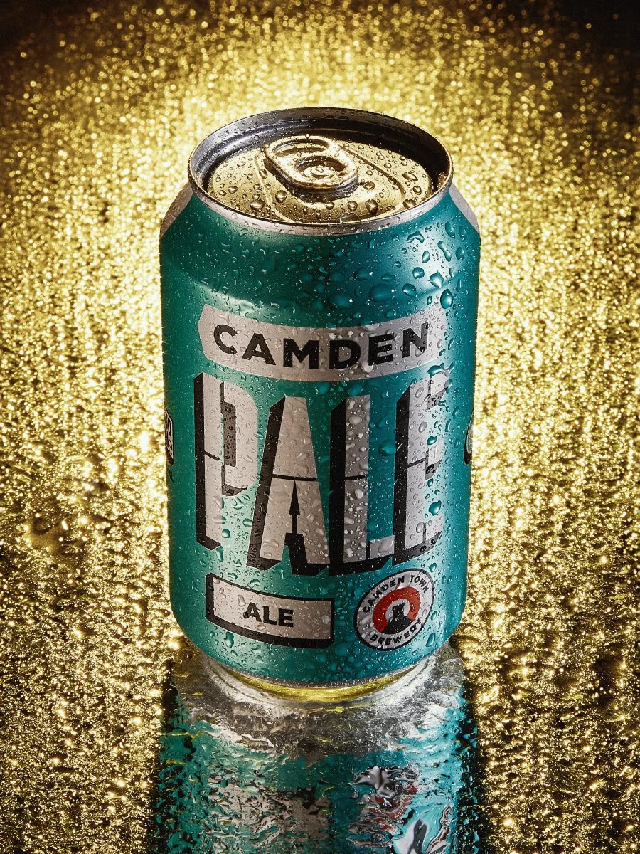 A teal Camden Pale Ale beer can with water droplets against a sparkling gold glitter background.