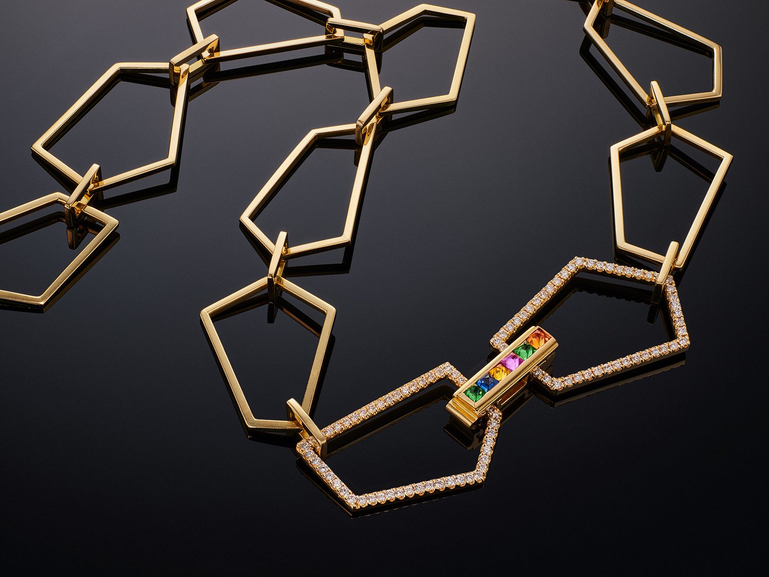 Gold necklace featuring geometric pentagon links with diamond accents and a multi-coloured rainbow gemstone bar.