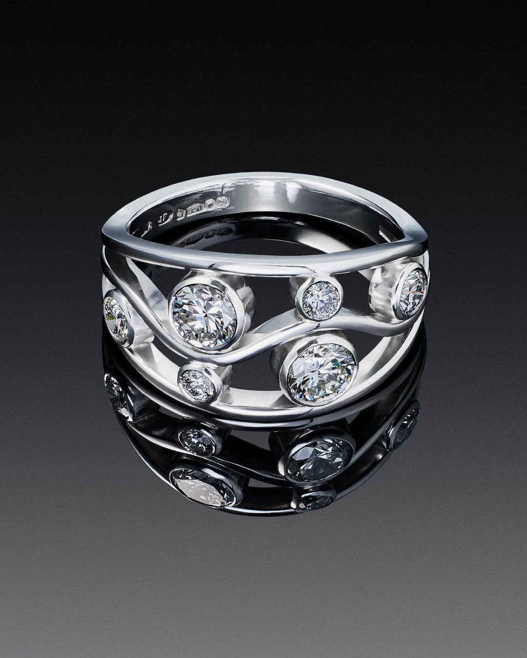 Wide silver band ring with round bezel-set diamonds in a wavy pattern, reflecting on a dark surface.